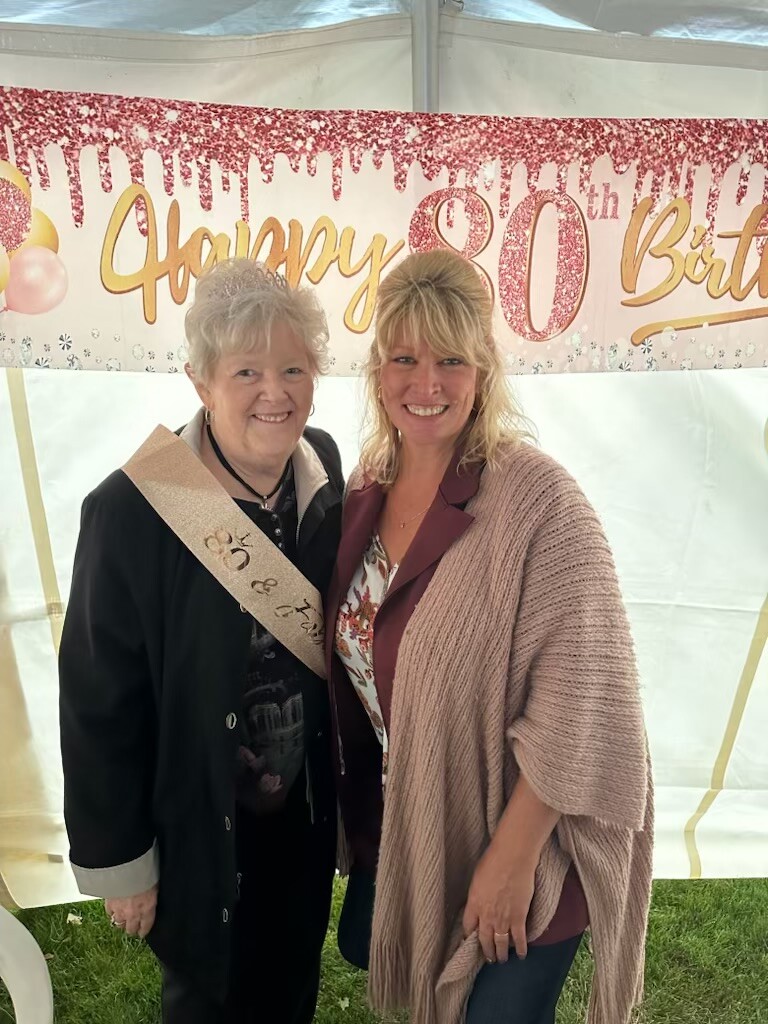 Two smiling women pose at an 80th birthday celebration under a pink glitter banner reading 'Happy 80th Birthday'.