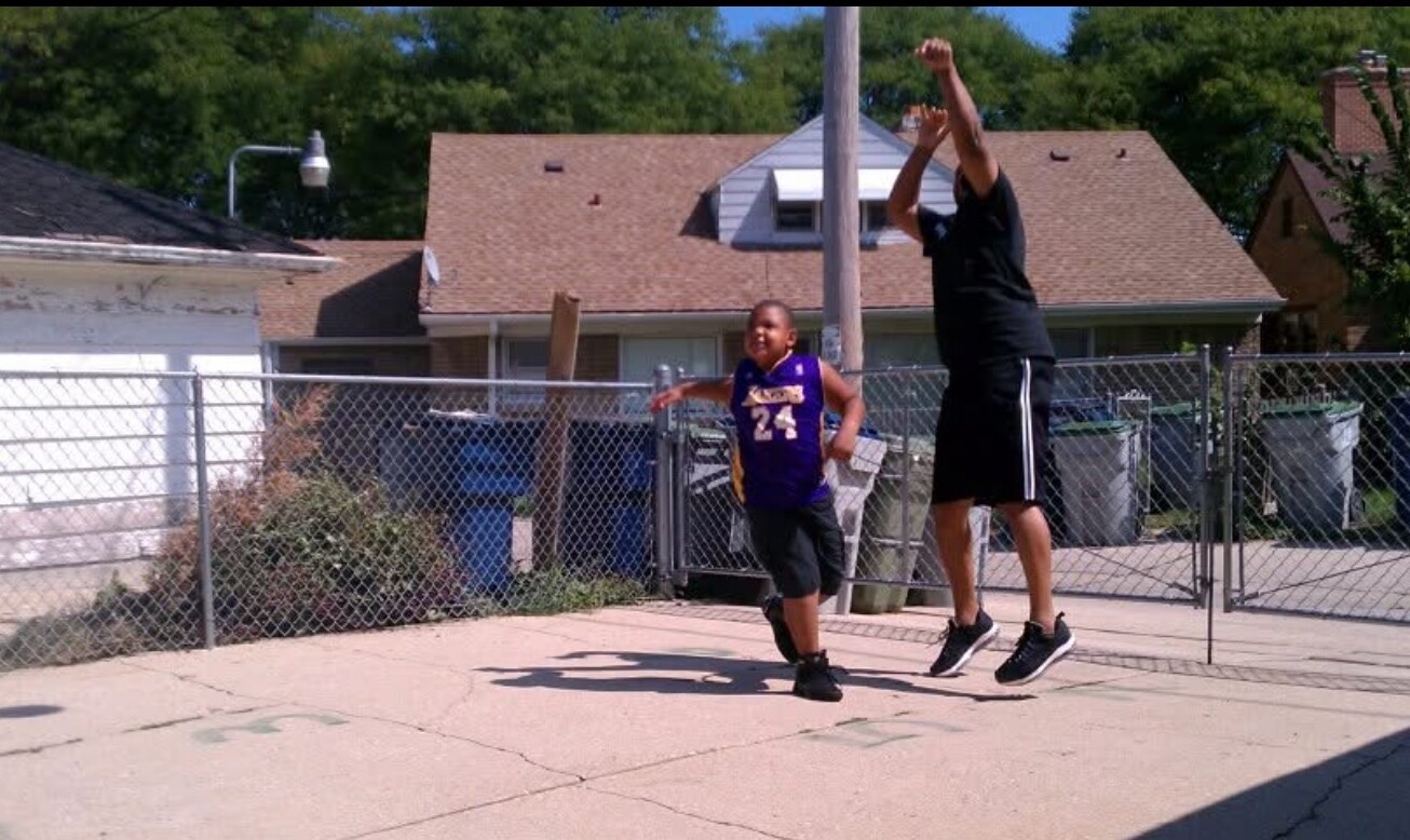 Man and boy playing basketball in a driveway, both mid-air as they contest a shot near a chain-link fence.