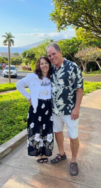 Smiling couple posing on a sunny sidewalk; man in a tropical shirt and light shorts, woman in a white cardigan and black floral skirt, with palm trees and hills in the background.