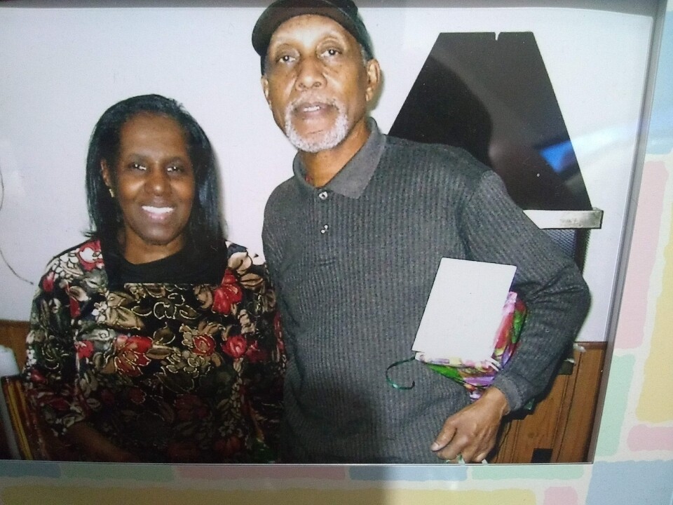 Smiling man and woman posing together indoors; the man holds a wrapped gift and a card.