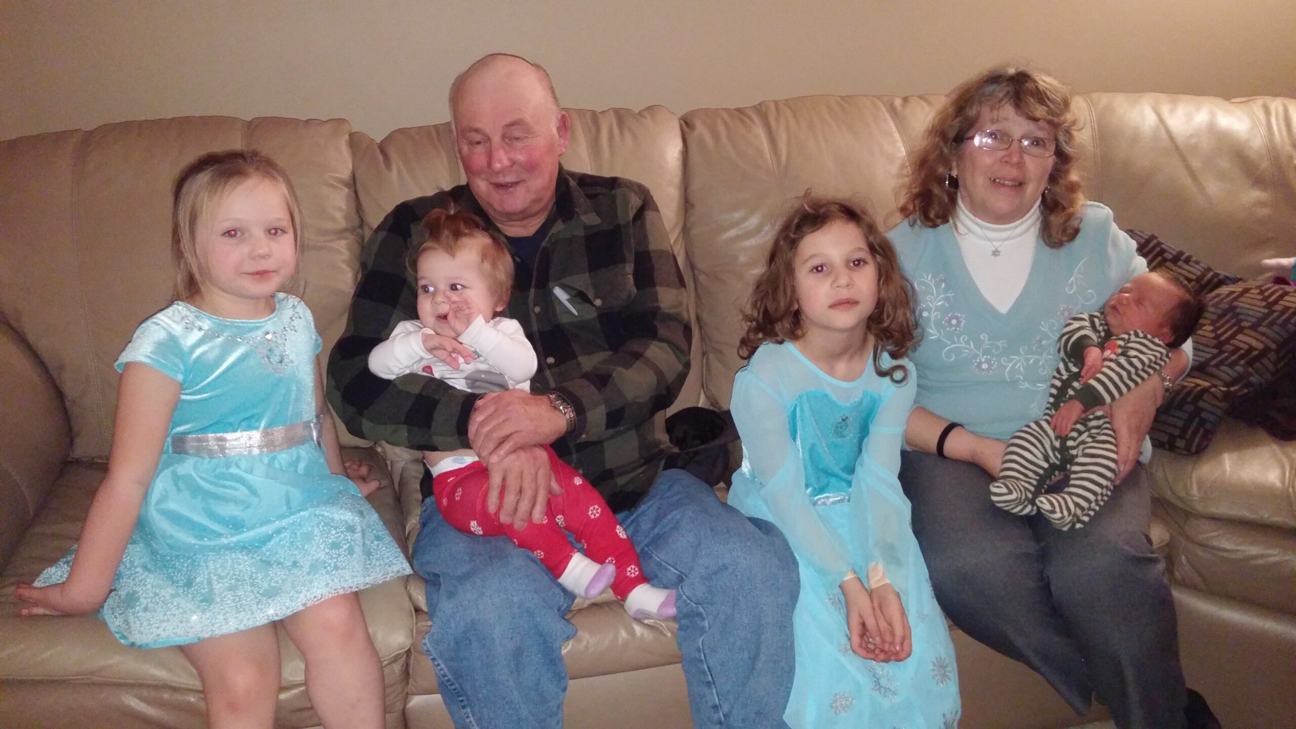 Grandfather and grandmother sit with three young girls on a beige couch, a baby in the grandfather's lap; family portrait in living room.
