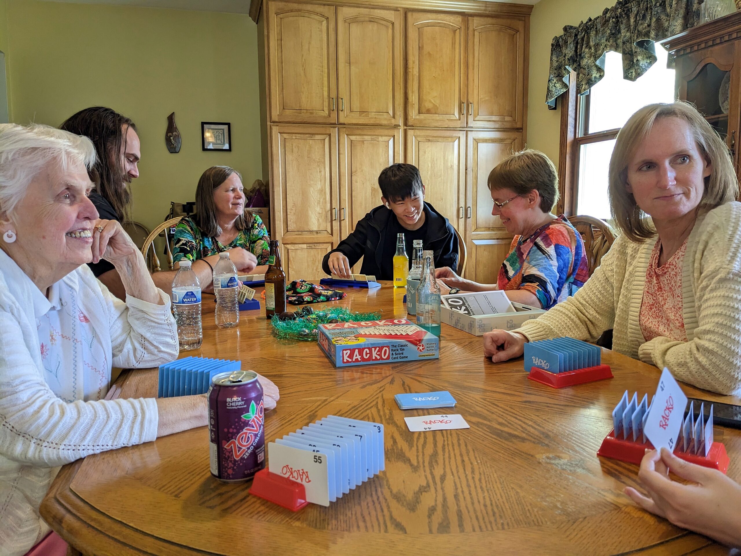 Six adults sit around a wooden dining table in a kitchen, playing a card game with blue racks and cards scattered among bottles and boards on the table.