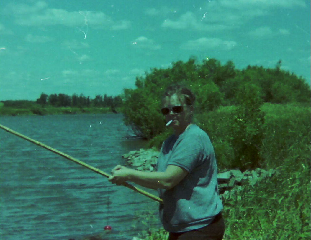 Man in a blue T-shirt and sunglasses fishes along a lakeside, cigarette in mouth, with green foliage in the background.
