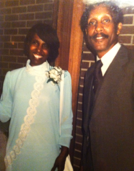 Two people posing for a photo at a formal event: a woman in a light blue dress with white lace and a corsage, and a man in a dark suit and striped tie, standing in front of a brick wall.