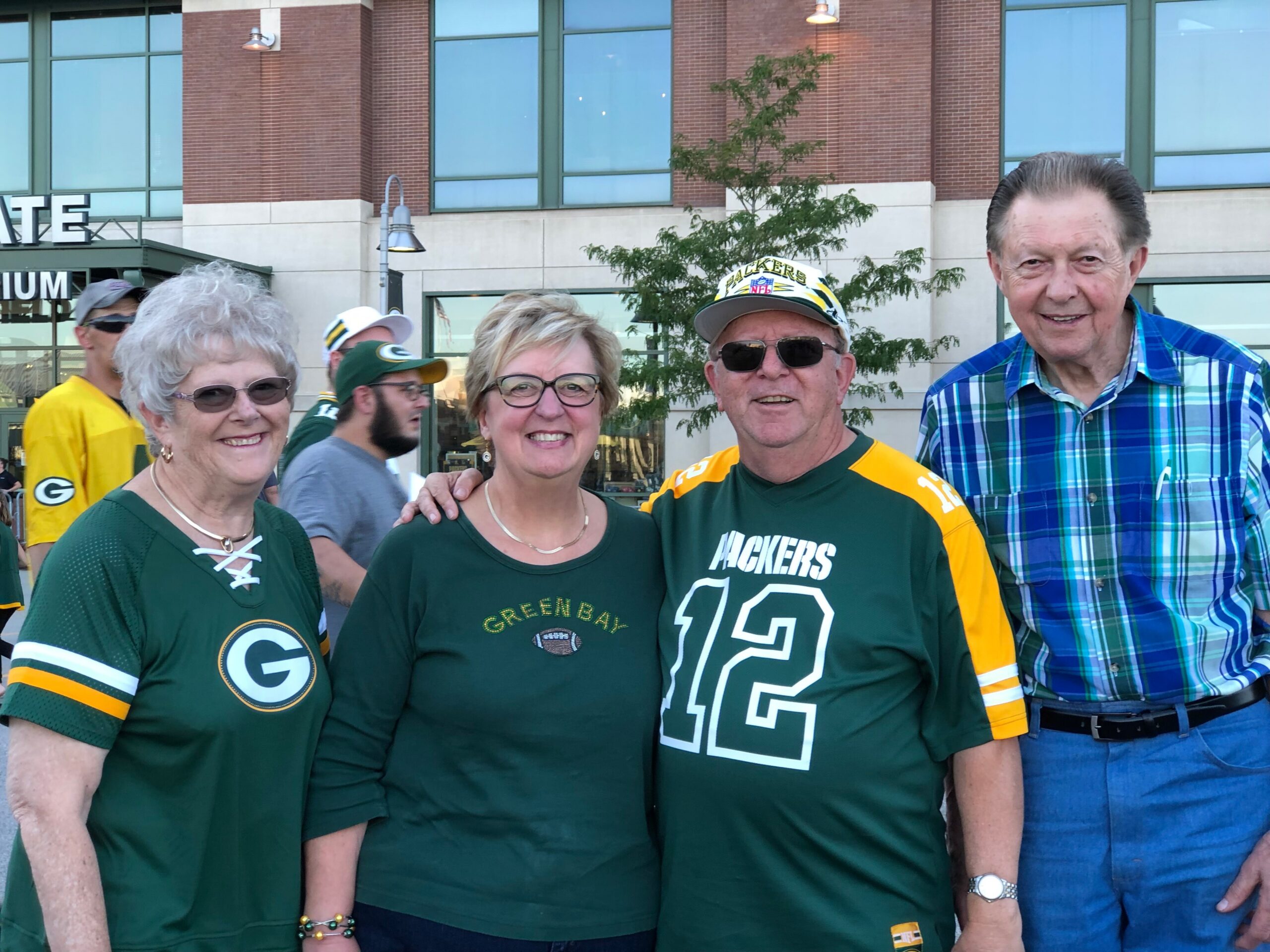 Smiling group of Packers fans in green and yellow jerseys stands outdoors in front of a brick building.