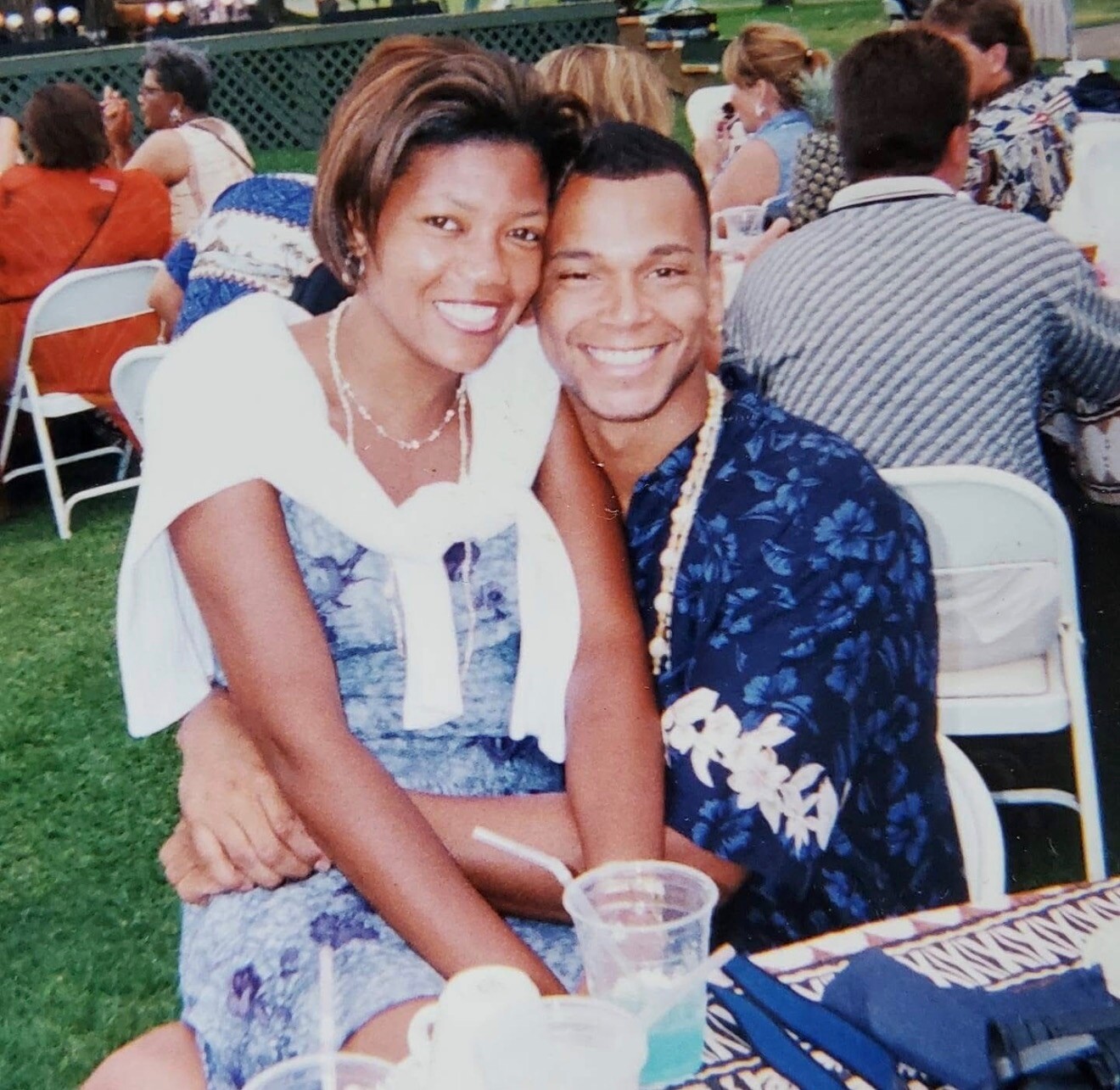 Smiling couple sitting close together at an outdoor event, wearing summery outfits and drinks on the table in front of them