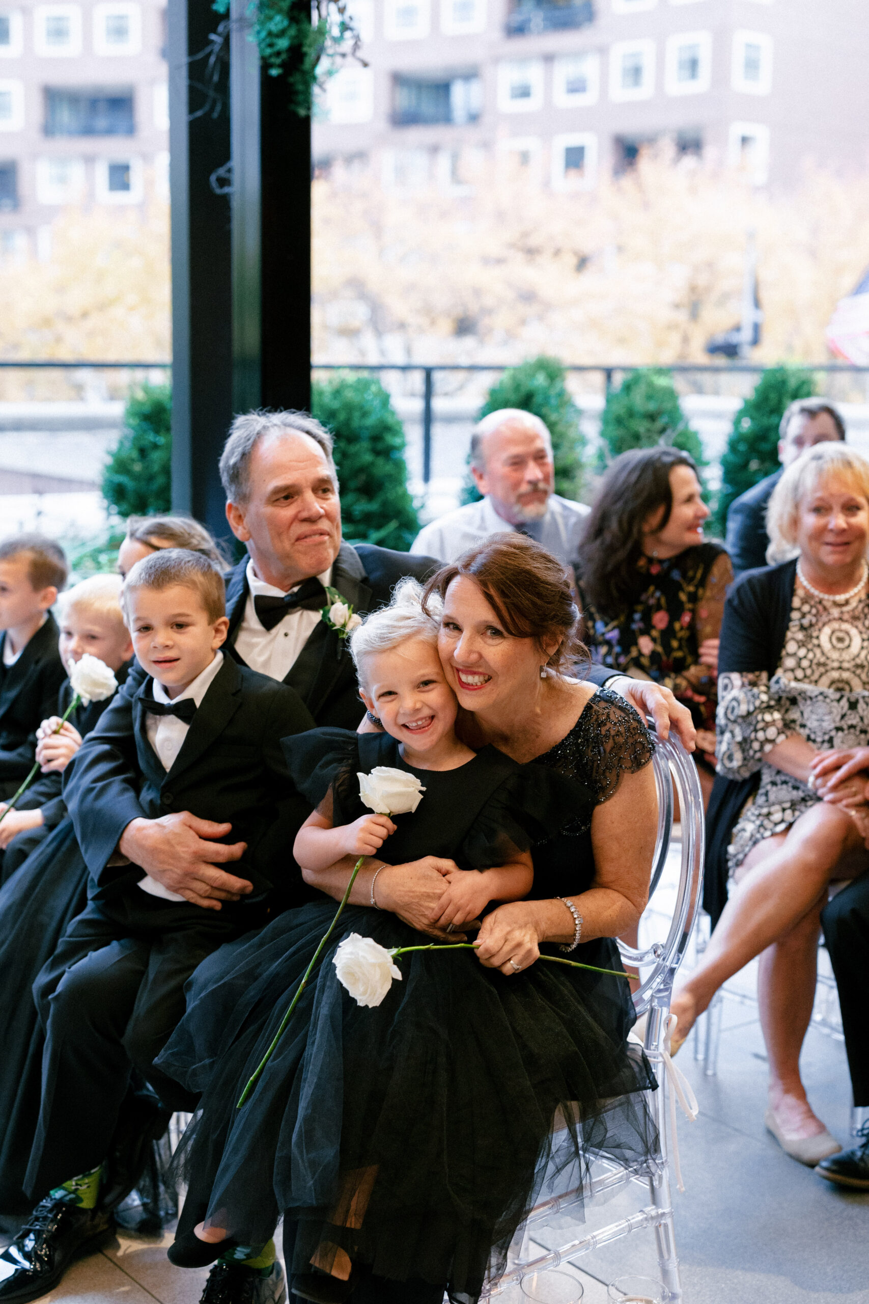 Smiling woman in a black lace dress sits with a young girl on her lap, both holding white roses at a formal event with guests nearby.