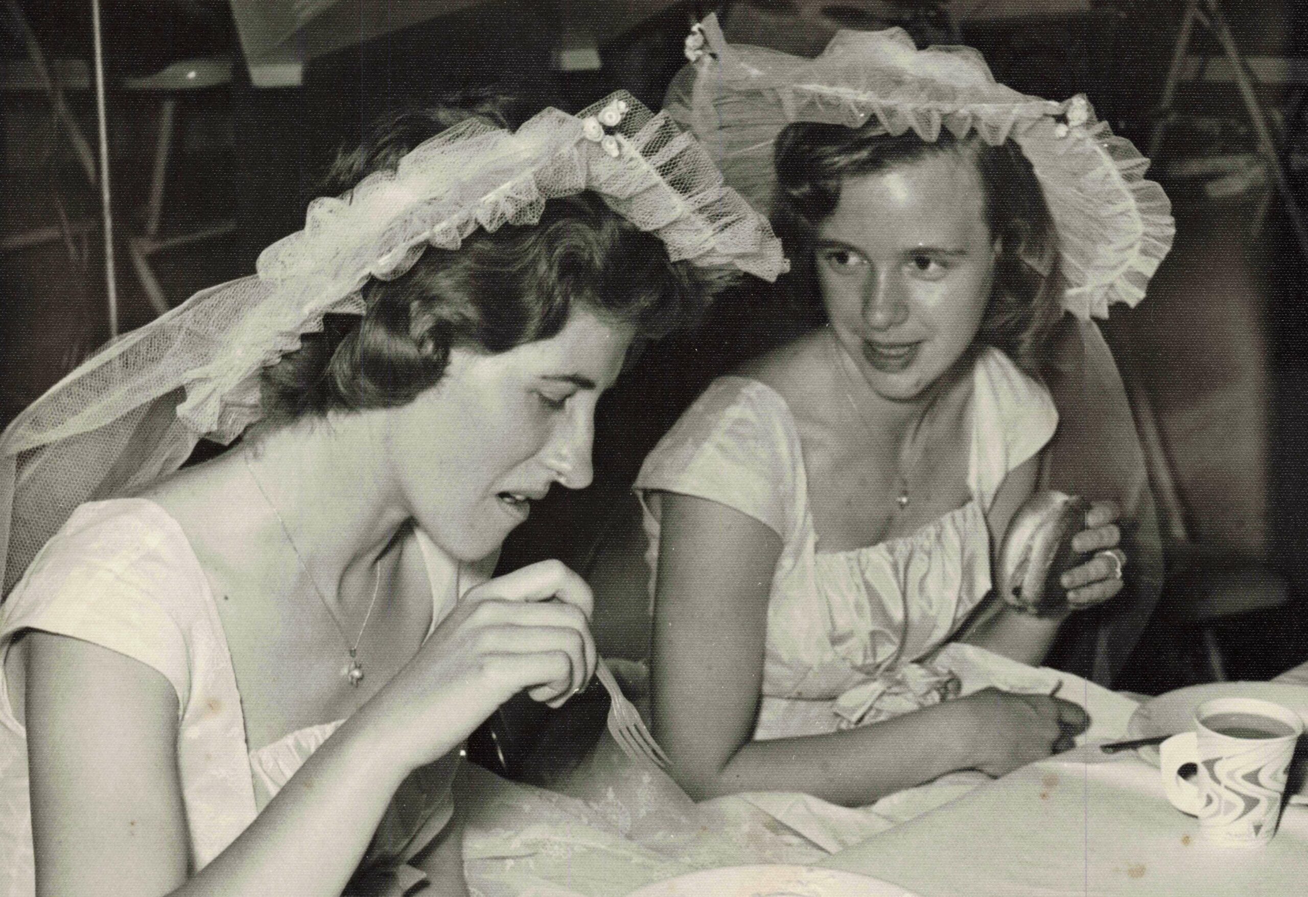 Two women wearing wedding veils sit at a table, cutting and sharing a slice of cake at a wedding reception.