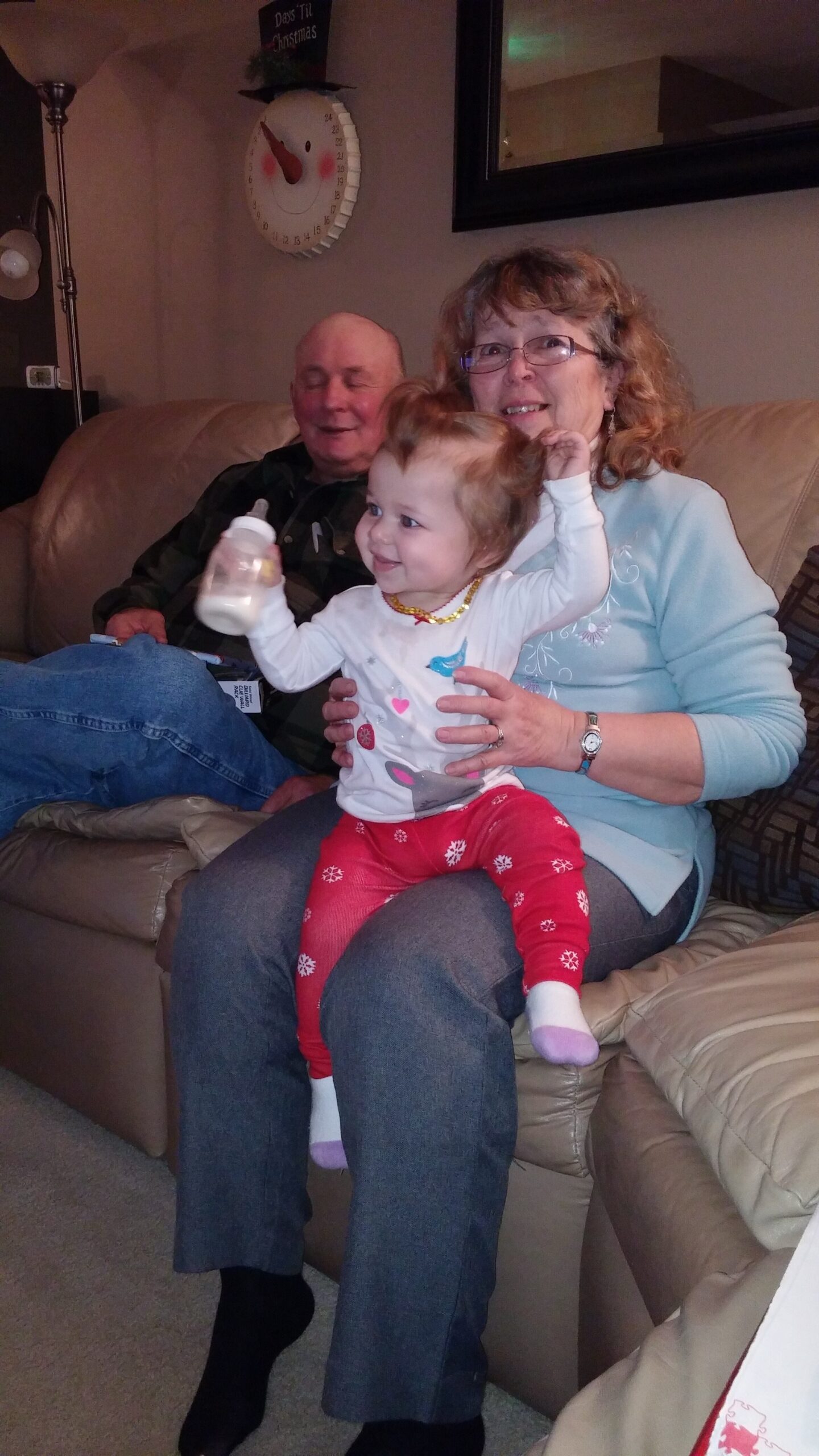 Young toddler on a grandma's lap raises a bottle while sitting on a beige couch; an older man watches in the background at home.