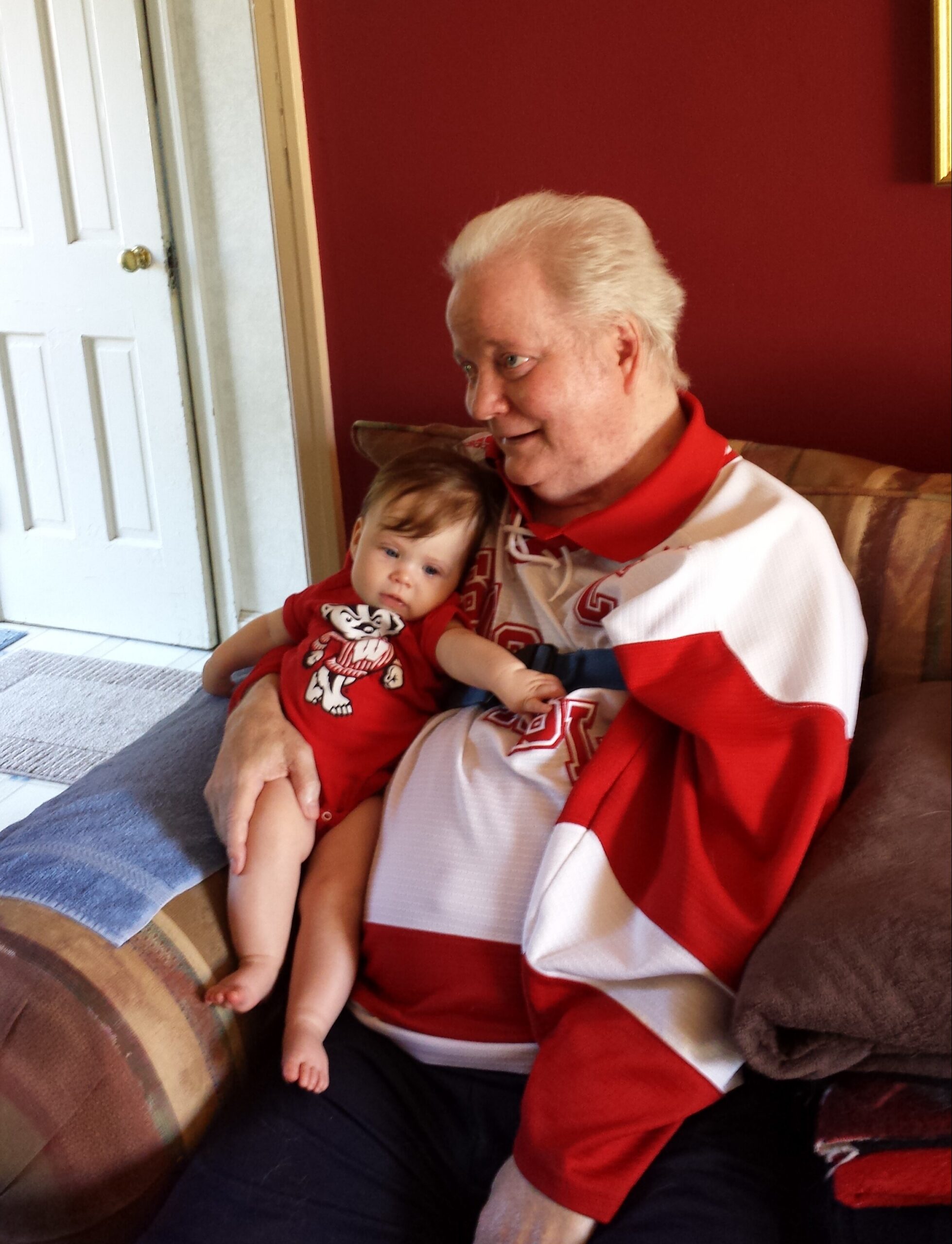 Older man with white hair wearing a red and white hockey jersey sits on a couch, holding a small baby in a red outfit.