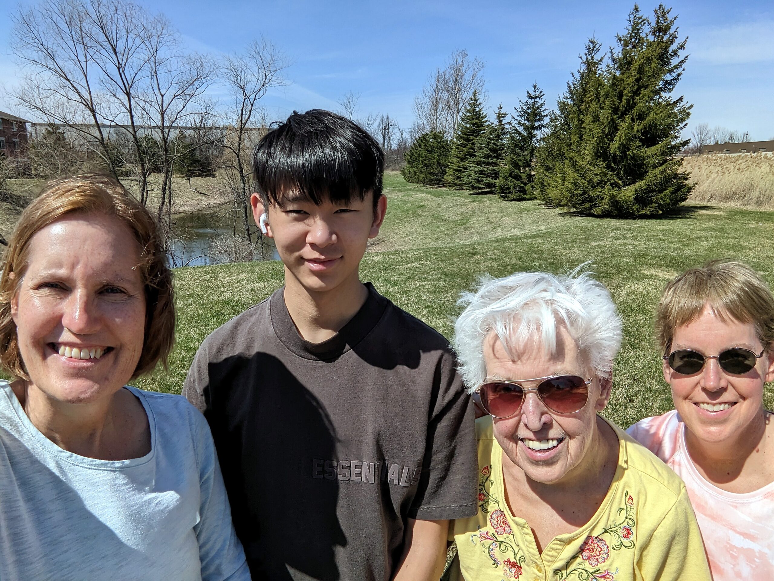 Smiling group photo taken outdoors in a park with bare and evergreen trees and a blue sky behind them, people posing for a selfie.