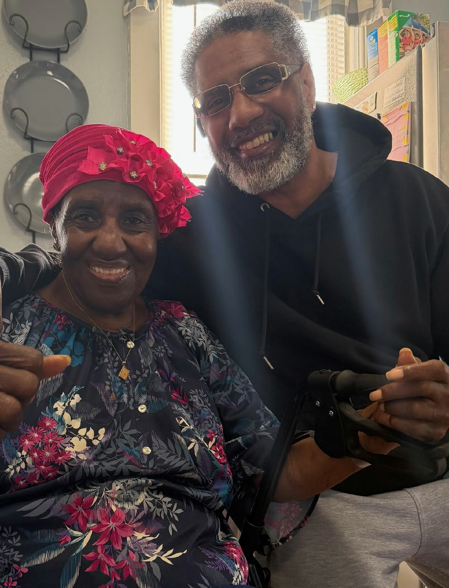 Smiling elderly woman in a bright pink headwrap and floral blouse beside a man with gray beard and glasses, posing together indoors.