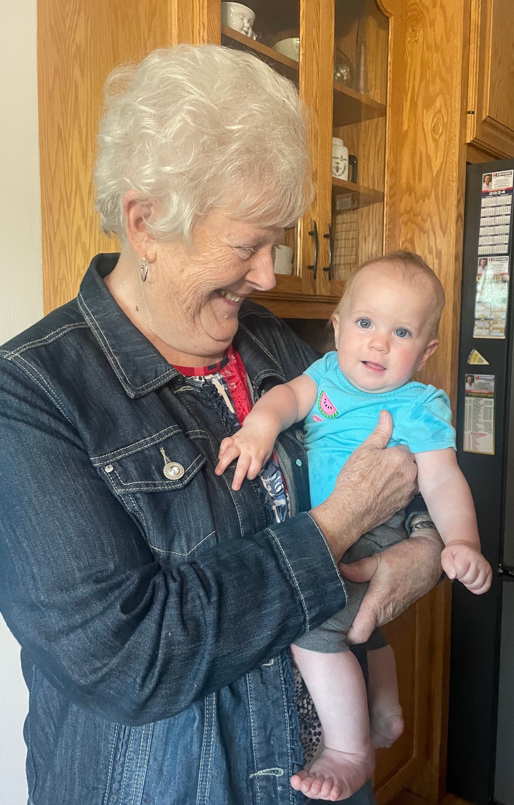 Smiling elderly woman holding a baby in a kitchen with wooden cabinets behind them, both looking at each other.