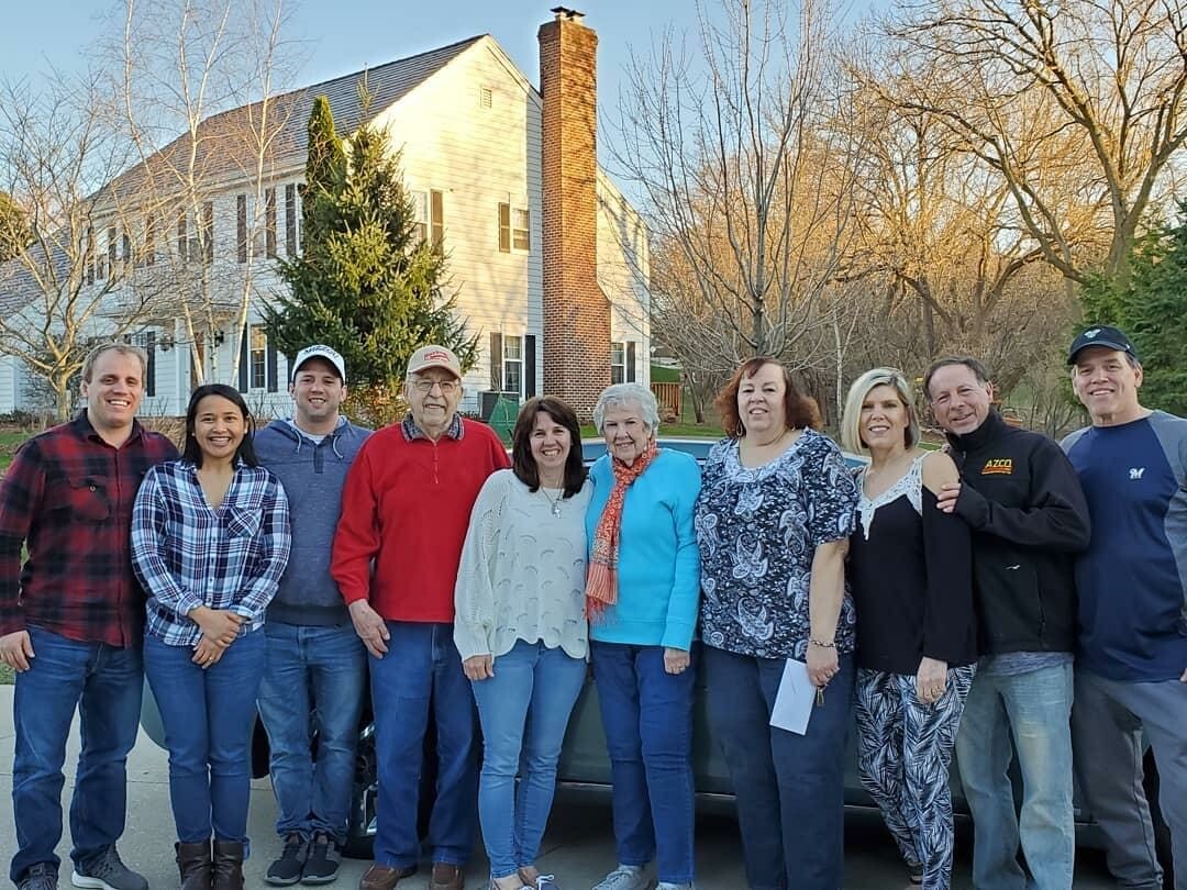 Eleven adults lined up for a group photo on a driveway in front of a two-story suburban house with a tall chimney.