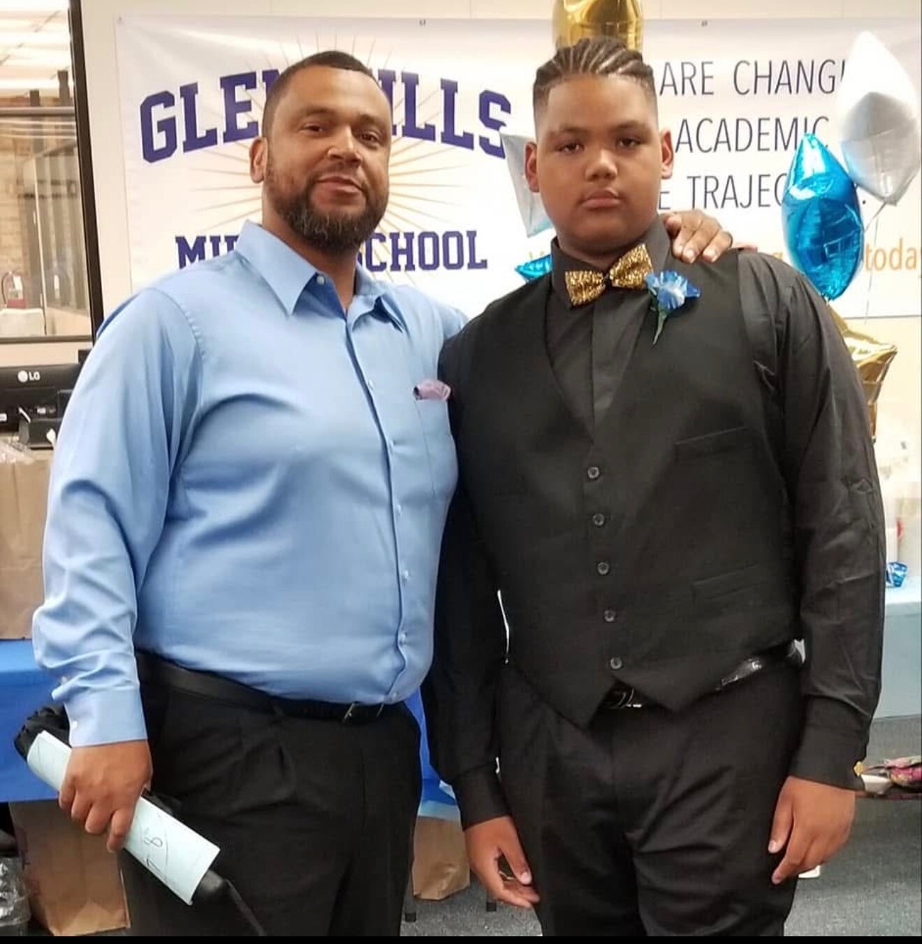 Two men posing together at a school event, one in a light blue shirt and the other in a black suit with a gold bow tie and blue boutonniere.