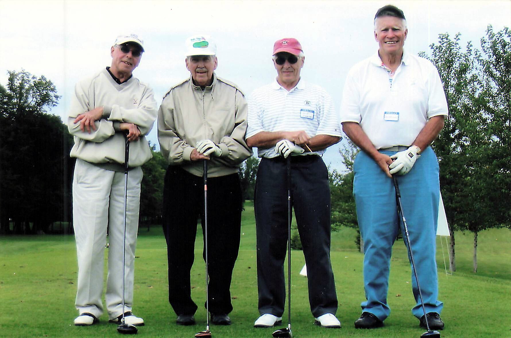 Four men posing with golf clubs on a green golf course, wearing hats and casual attire.