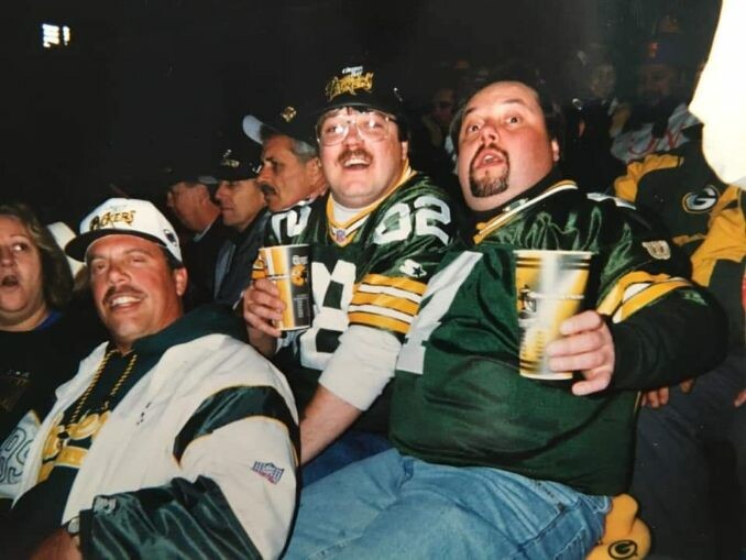 Group of Green Bay Packers fans at a game, smiling and holding beer cups in stadium seating.