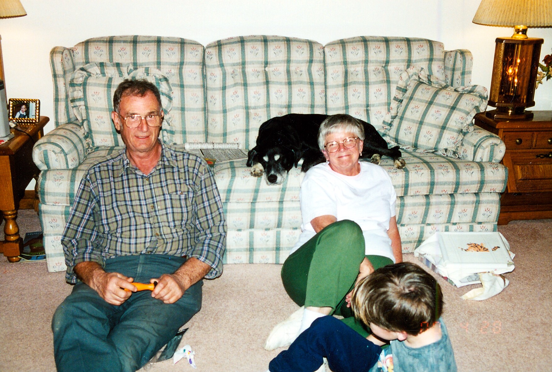 An elderly man and woman sit on a plaid sofa with a black dog resting between them in a living room.