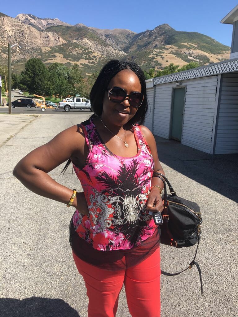 Casual portrait of a woman in a pink tropical-print top and red pants, wearing sunglasses, standing outdoors with mountains in the background, holding a car key fob beside a black bag.