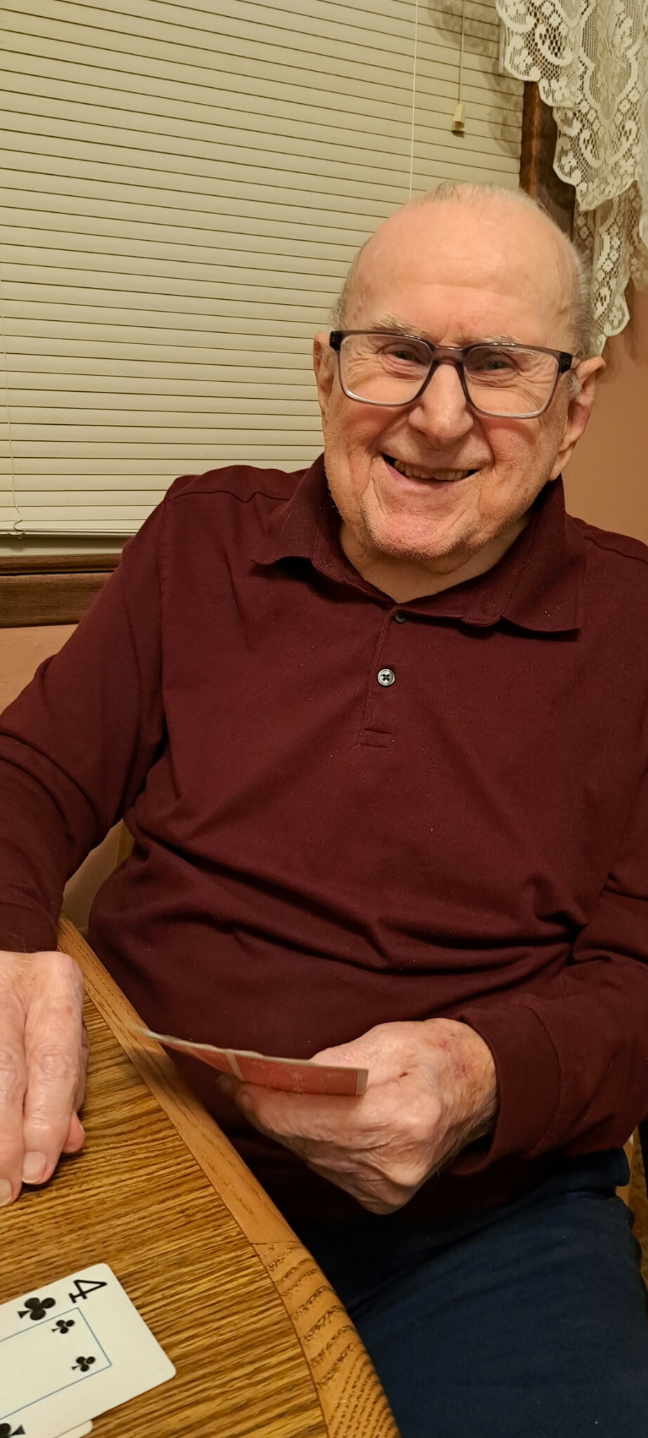 Smiling elderly man wearing a maroon polo and glasses, seated at a wooden table with playing cards in his hand.