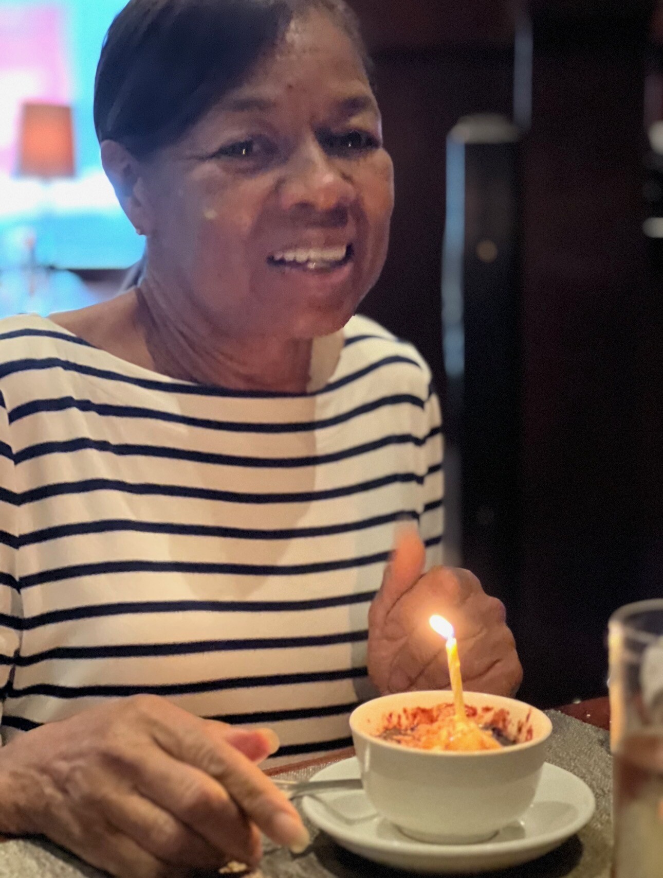 Smiling elderly woman in a striped shirt at a birthday cake with a lit candle in front of her.