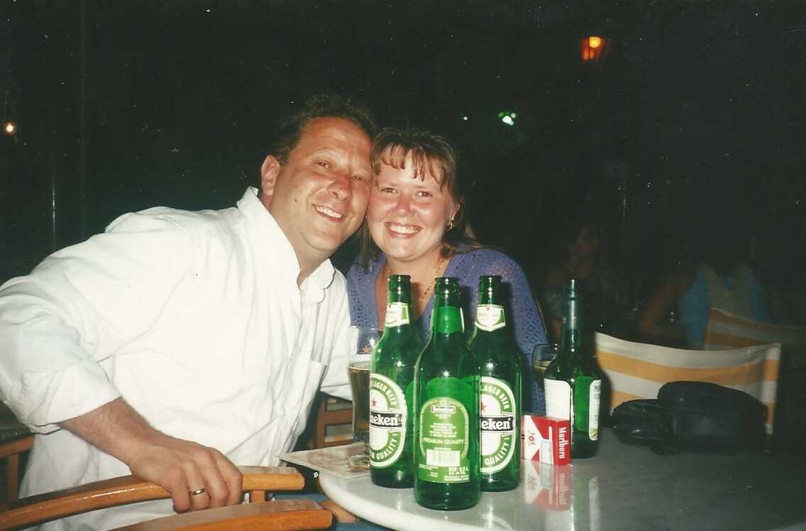 Smiling man and woman sit at a table with several green Heineken bottles at night outside.
