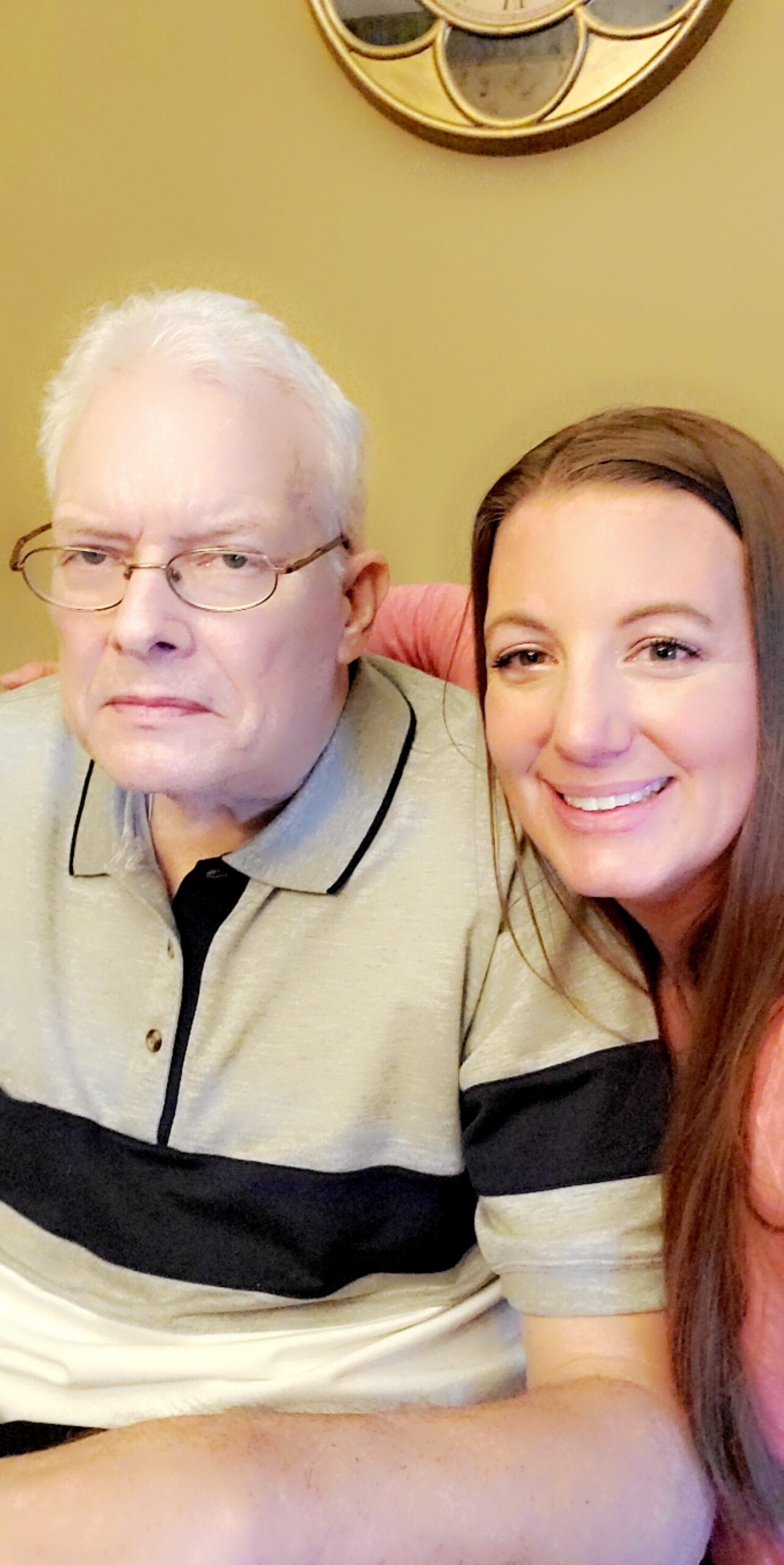 An older man with white hair and glasses sitting beside a smiling younger woman taking a selfie together in a warm room