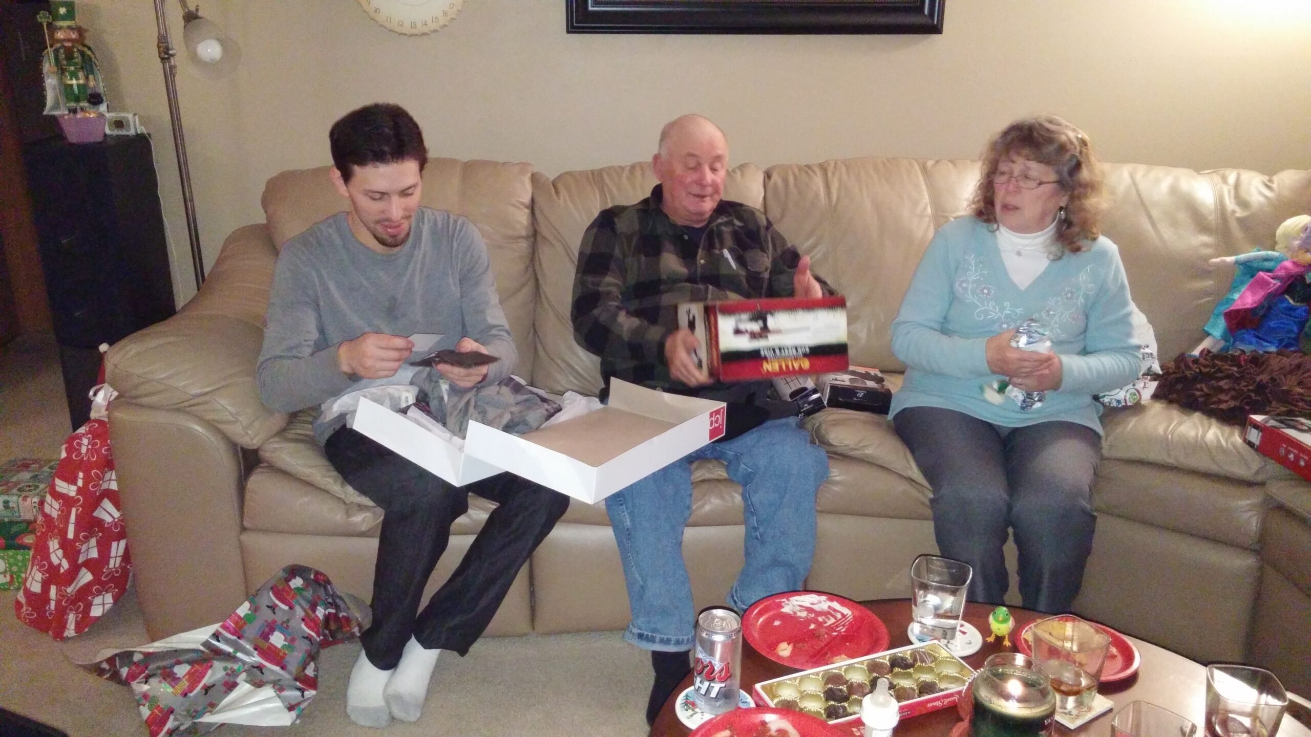 People on a beige sofa opening presents during a cozy holiday gathering with wrapping paper and gifts around them.