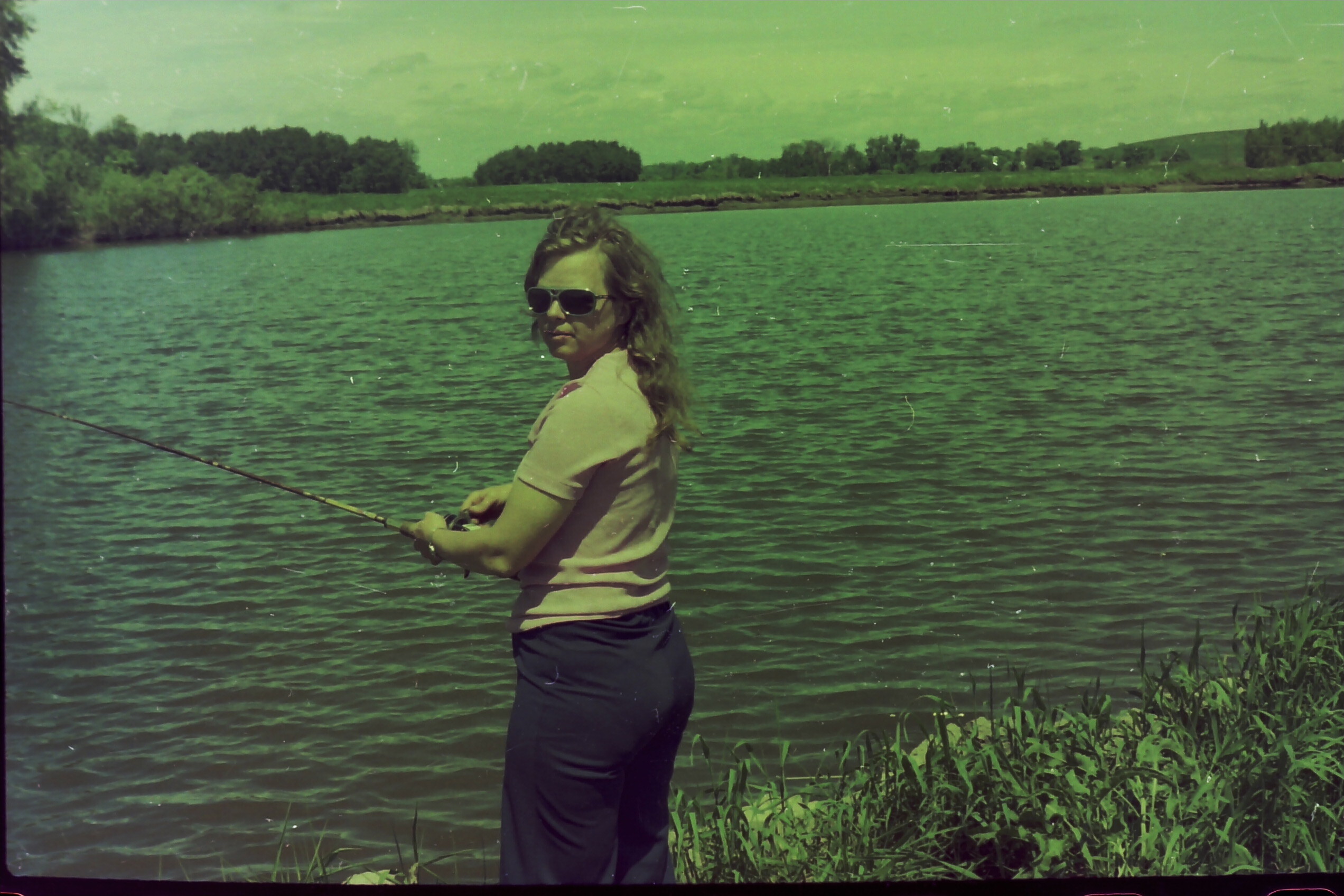 Woman fishing by a lake in a pink shirt, wearing sunglasses, with reeds along the shore in the foreground.