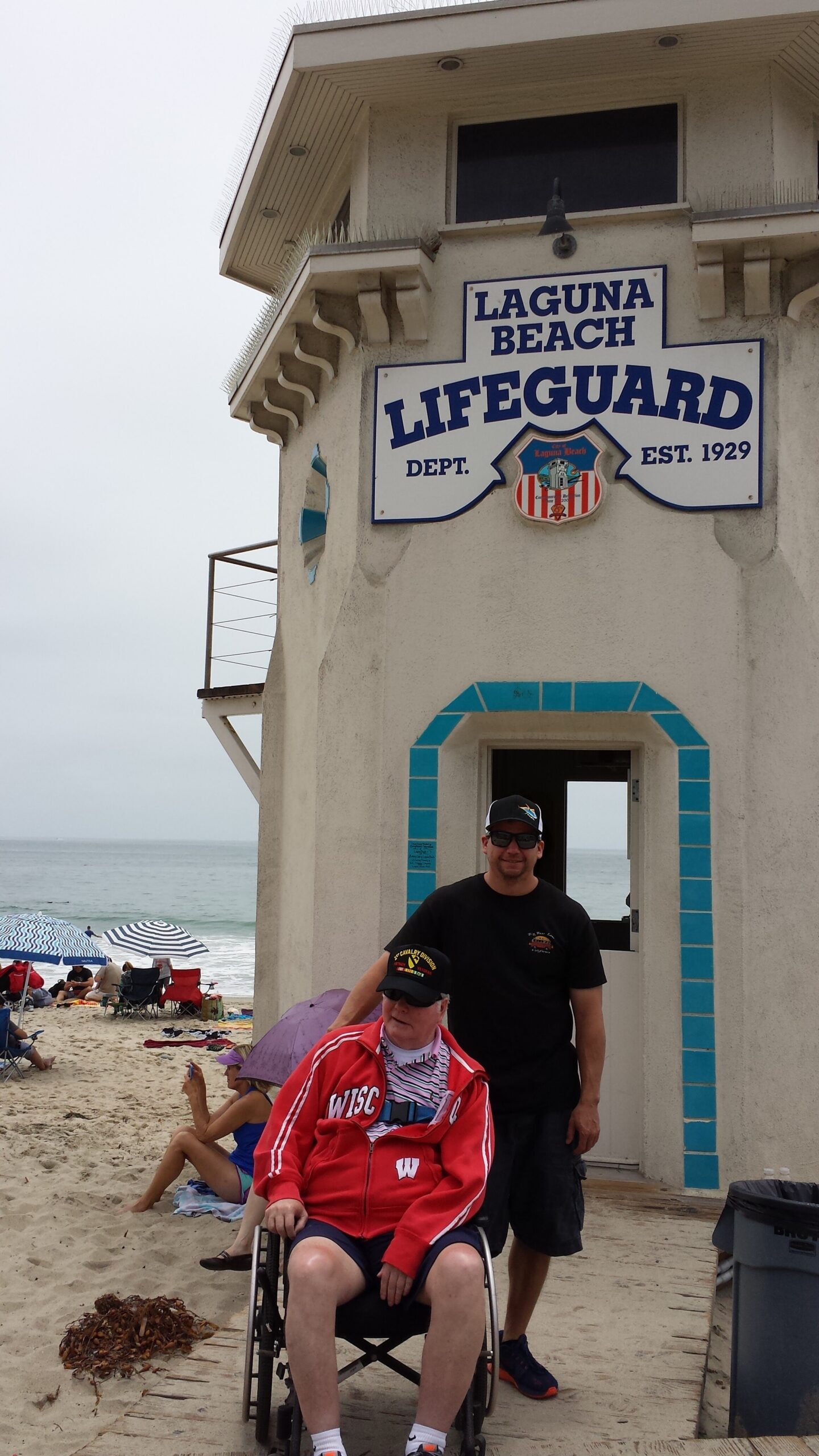 Lifeguard station with Laguna Beach sign on a building beside the beach; man in a wheelchair in front and others on the sand nearby under umbrellas.