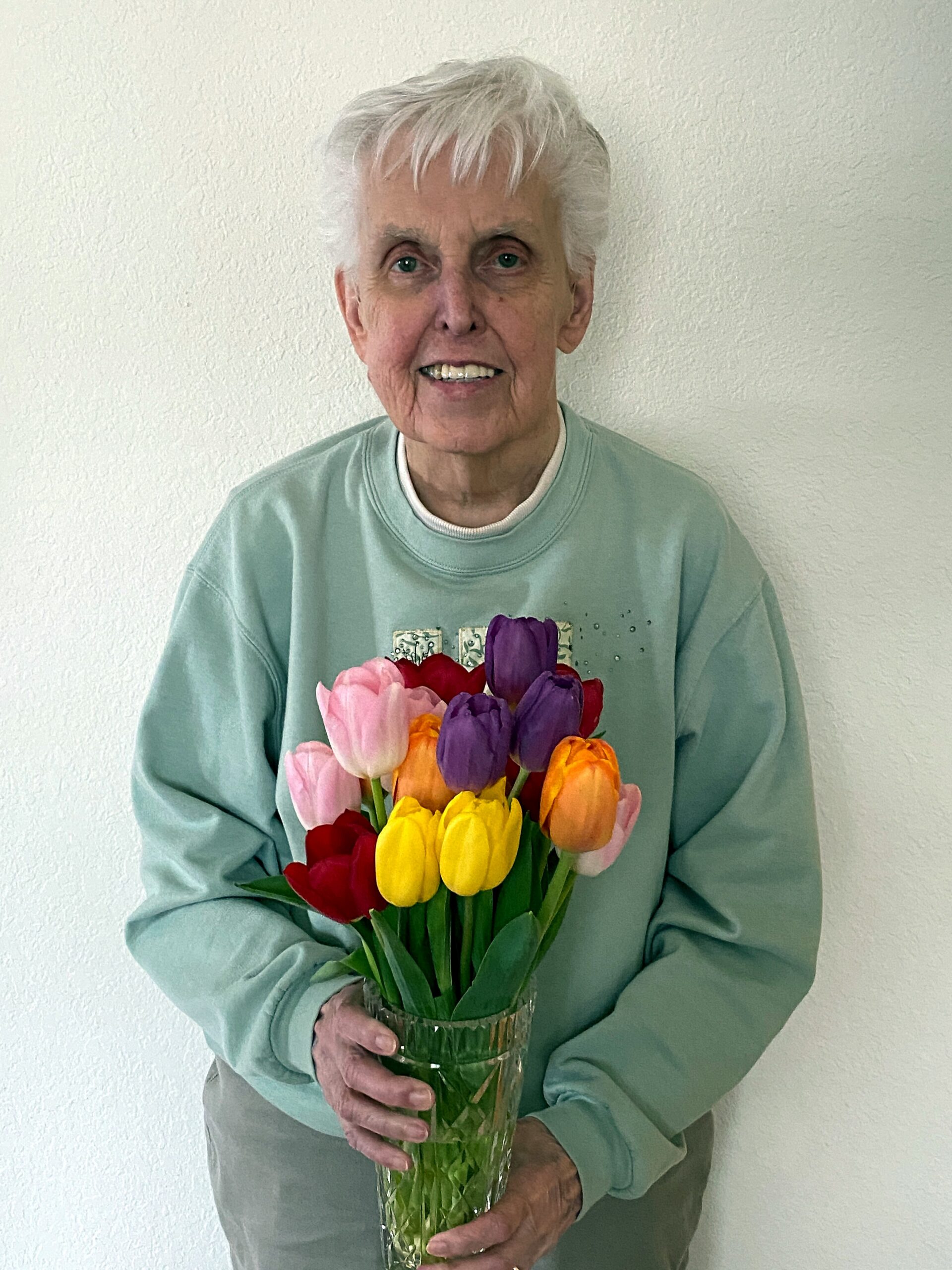 Older person with short white hair in a light green sweatshirt, smiling while holding a vase of mixed tulips (pink, red, purple, yellow).
