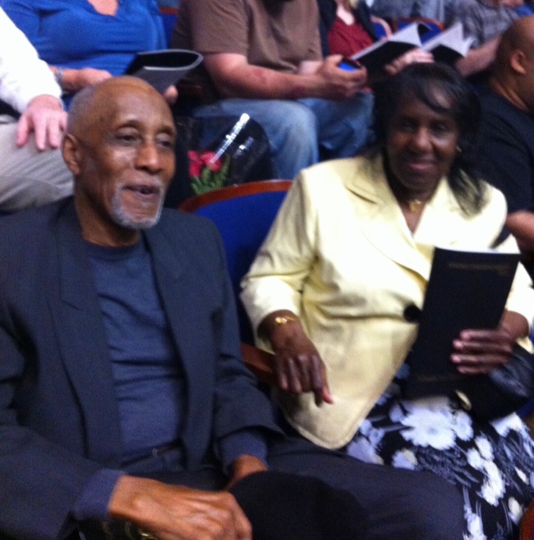 Senior man in a dark blazer and blue shirt sits beside a woman in a pale yellow blazer, holding a program at a crowded event venue.
