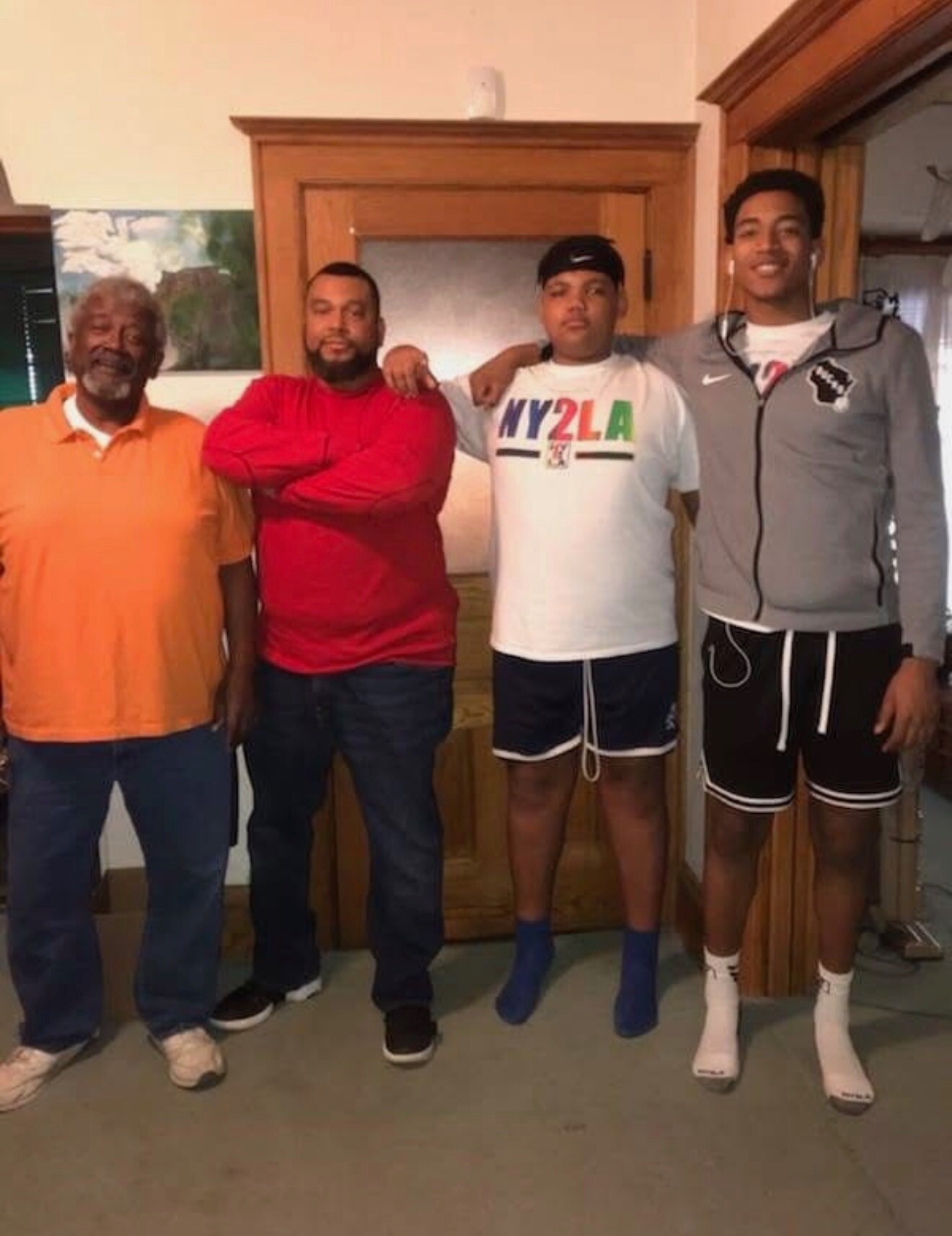 Four men pose together indoors, arms around each other, in a living room doorway setting; diverse ages, casual athletic wear.