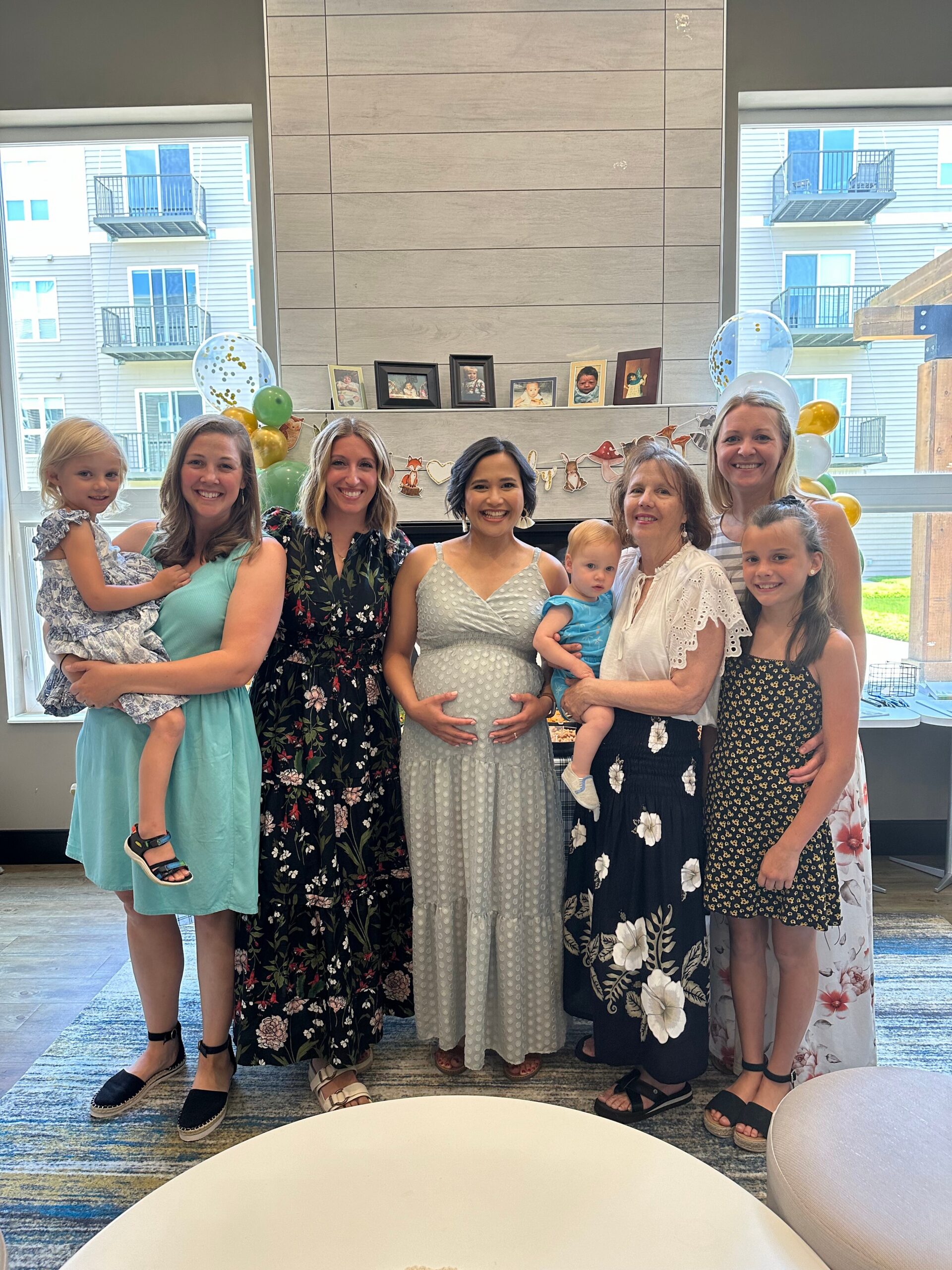 Group of women and children posing indoors for a baby shower or celebration around a pregnant woman in a light dress.