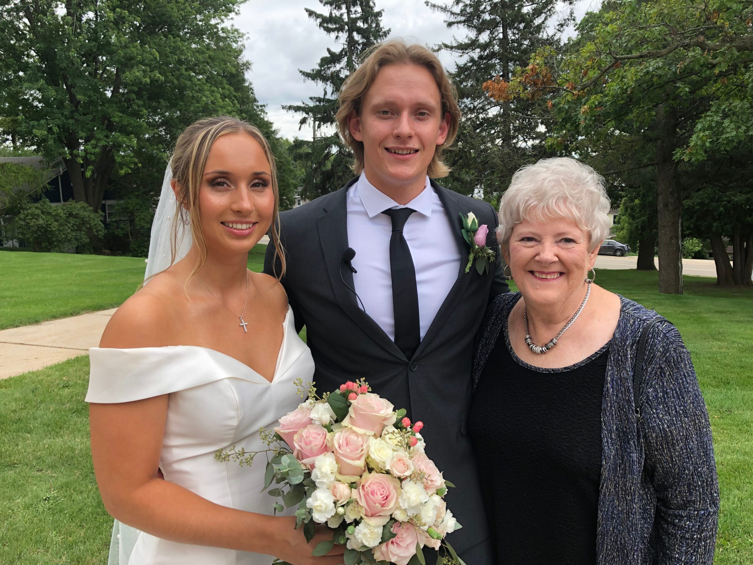 Bride in a white off‑the‑shoulder dress holding a pink bouquet, standing with a groom in a suit and an elderly woman outdoors at a wedding park.