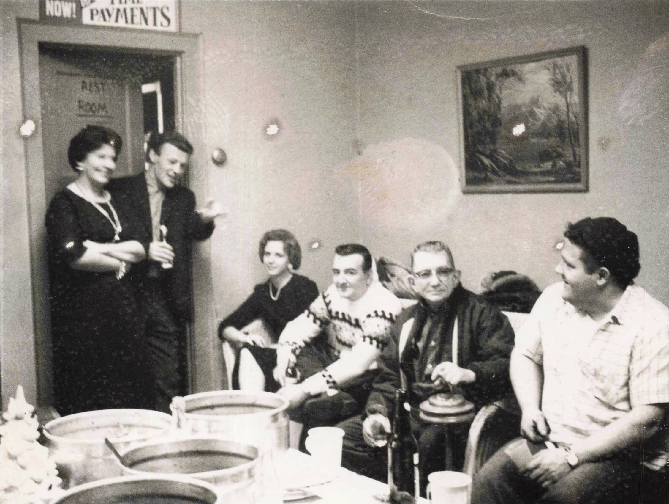 Black-and-white photo of coworkers at a mid-century office party, seated around a long table with drinks, while two women stand by the doorway.
