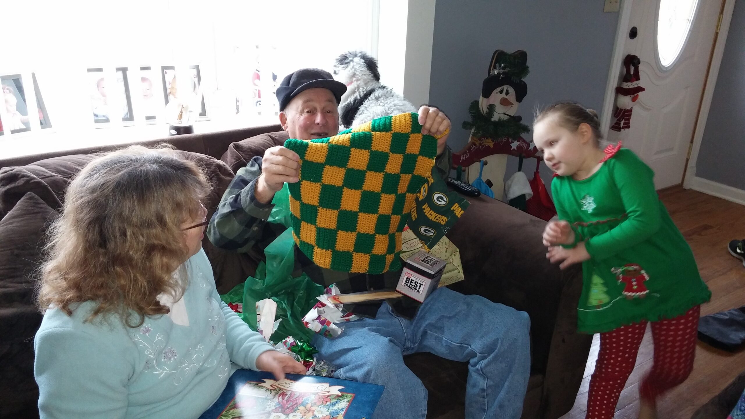 An older man on a couch proudly shows a green-and-yellow checkered knitted blanket to a smiling girl in a festive green outfit amid wrapping paper and gifts.
