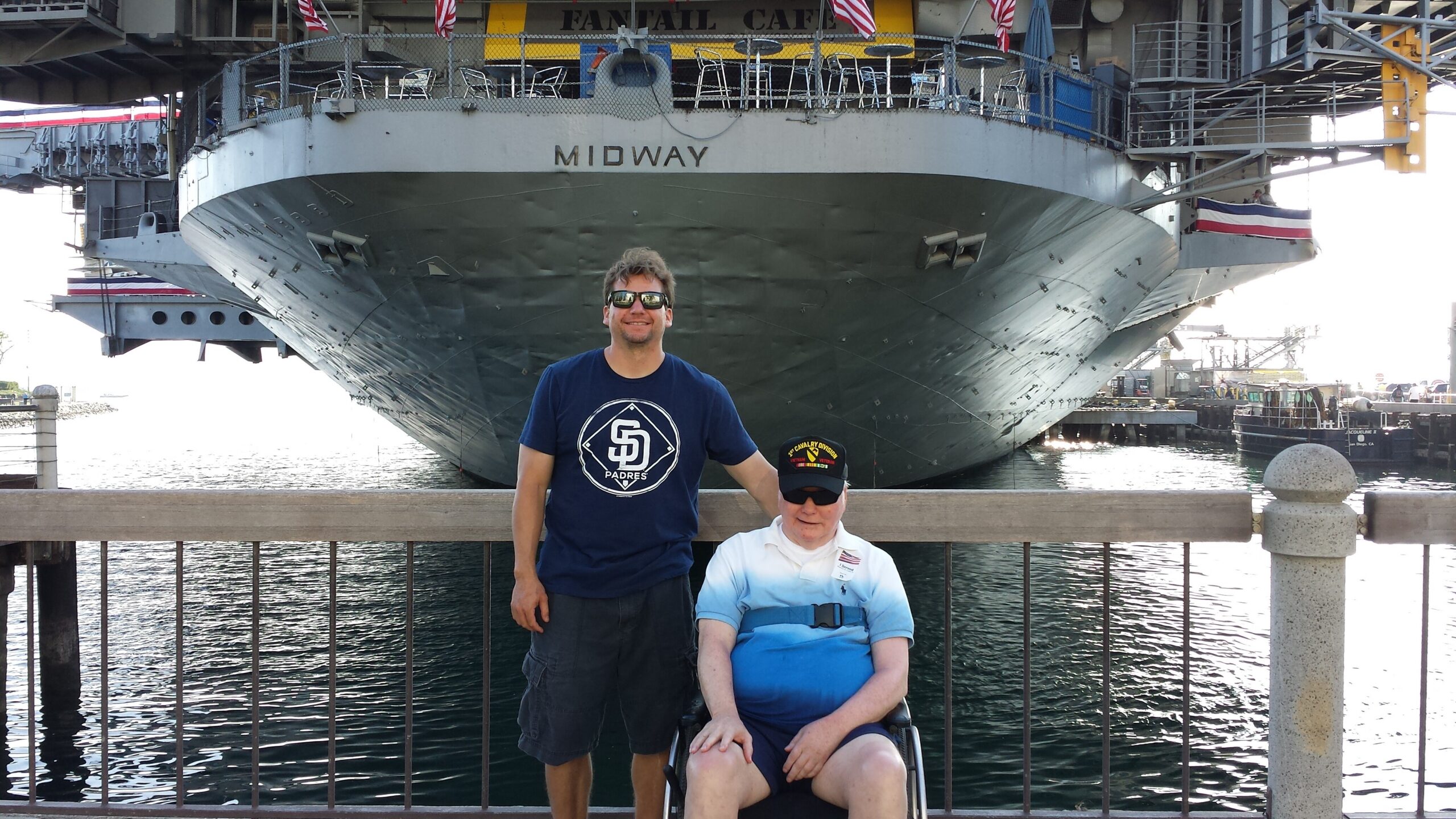 Two men pose in front of the large Midway aircraft carrier at a harbor, one standing and one in a wheelchair near the railing.