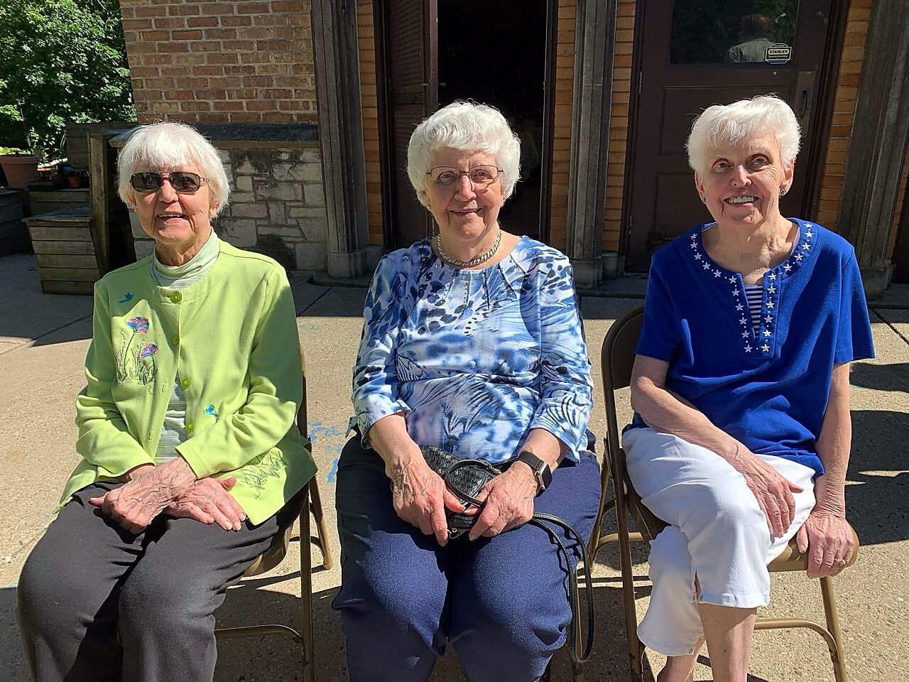 Three elderly women sit on folding chairs outside a brick building, smiling at the camera. The left wears a lime green jacket, the middle a blue patterned blouse, and the right a blue top with white stars.