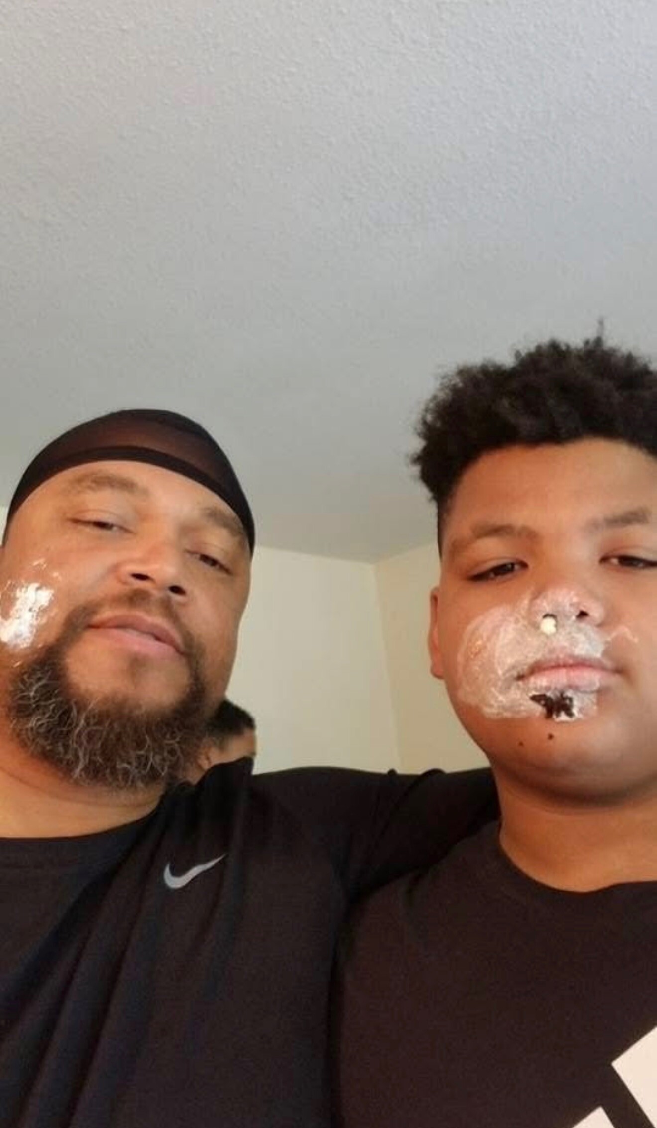 Two men take a casual selfie indoors; the man on the left wears a black durag and has a beard, the man on the right has white facial cream on his nose and cheeks.