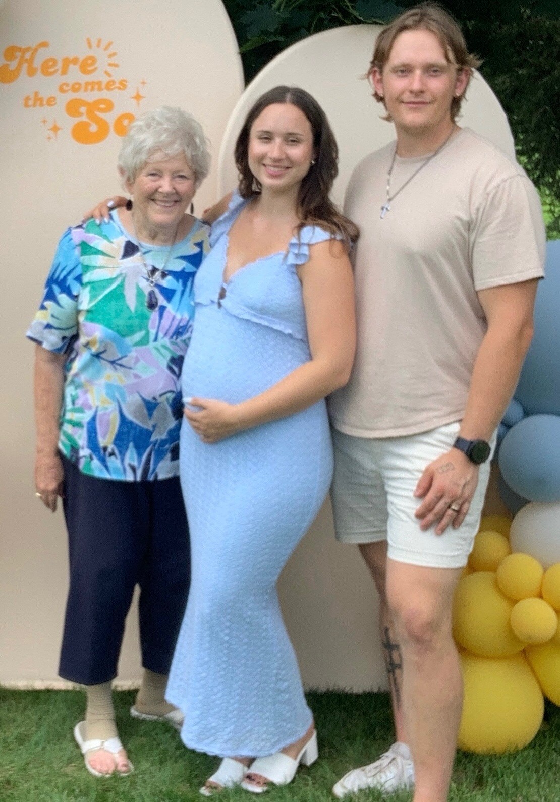 Three people posing outdoors: an older woman in a colorful top, a pregnant woman in a light blue dress, and a man in a beige T-shirt and shorts, with balloons in the background.