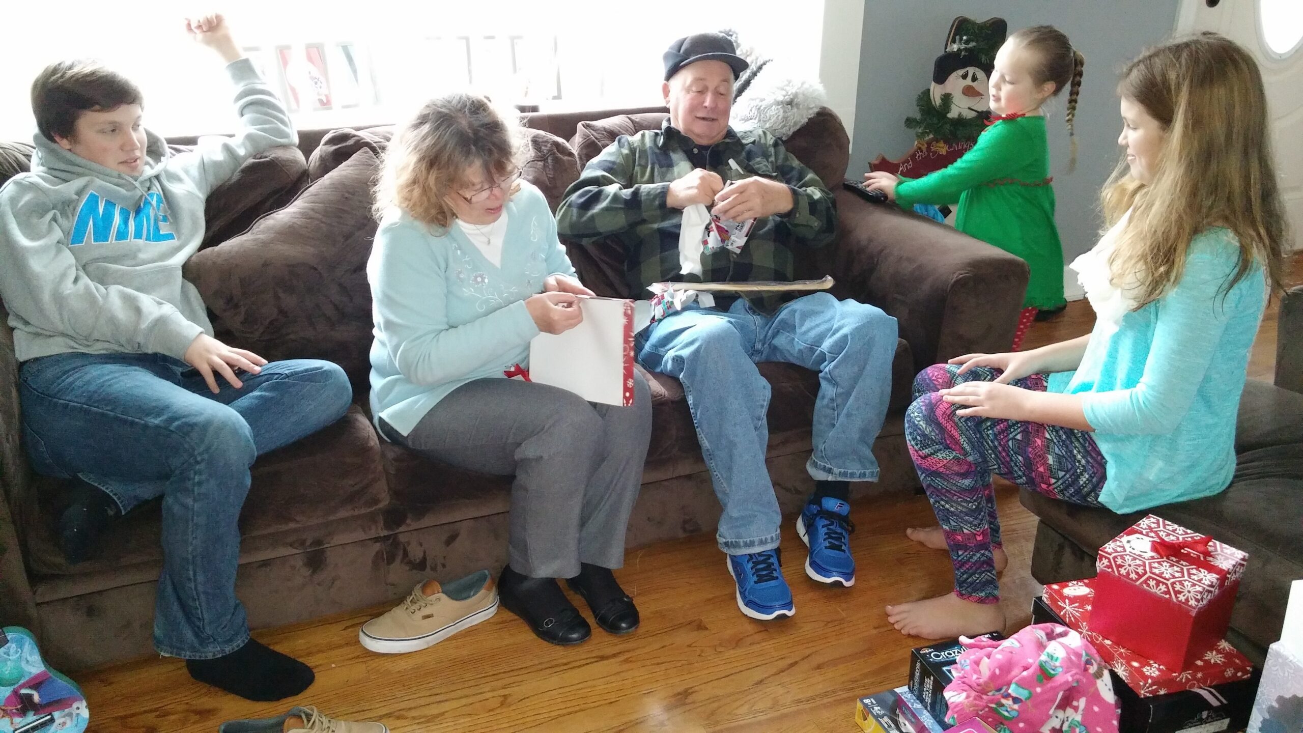 Family gathers in a living room for gift opening: two women and three children sit around a brown couch, with wrapped presents on the floor as an older man unpacks a gift in the center.