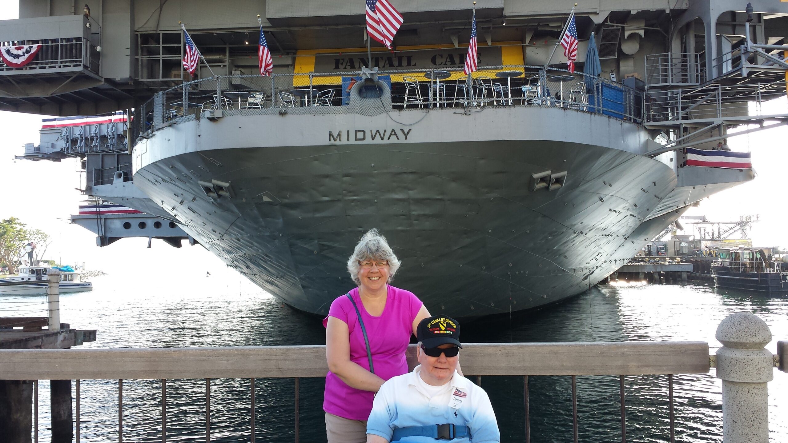 Two people pose at a railing in front of the USS Midway aircraft carrier, with the bow labeled MIDWAY and American flags flying overhead in the harbor setting.