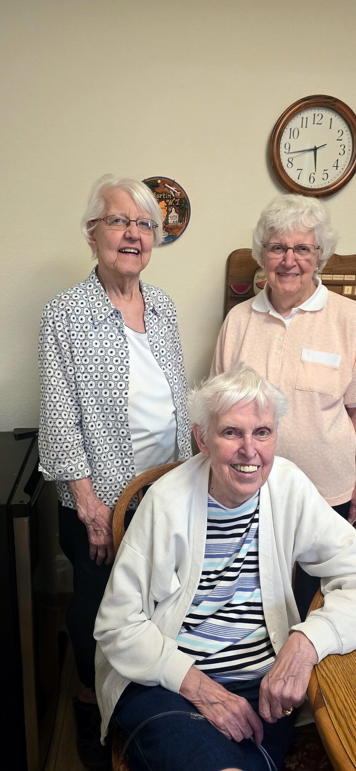 Three elderly women pose together indoors, one seated at a table smiling, two standing behind them with a clock on the wall.