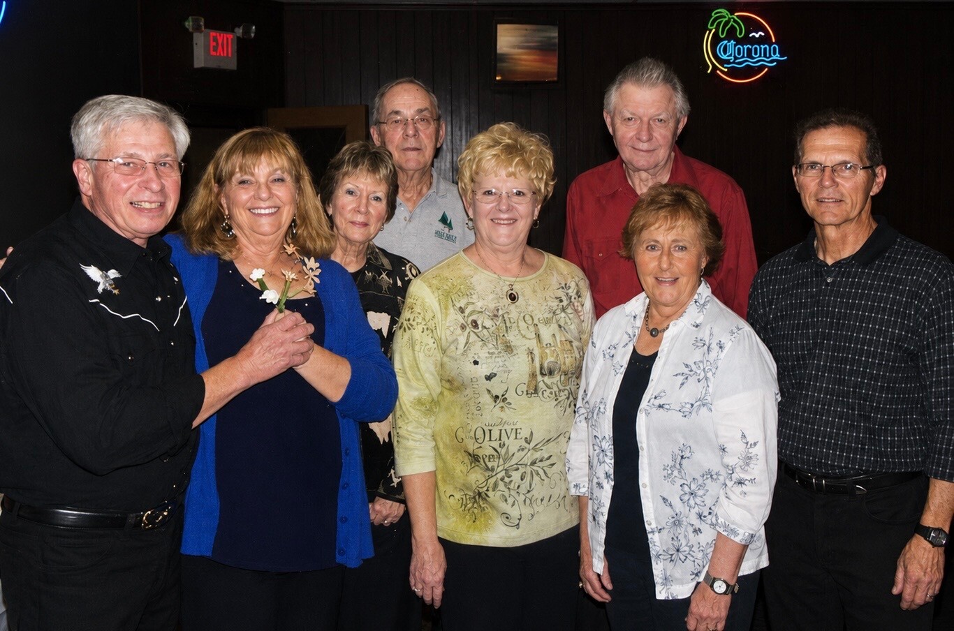 Group of eight adults posing for a photo in a dim bar, neon Corona sign visible in background, everyone smiling.