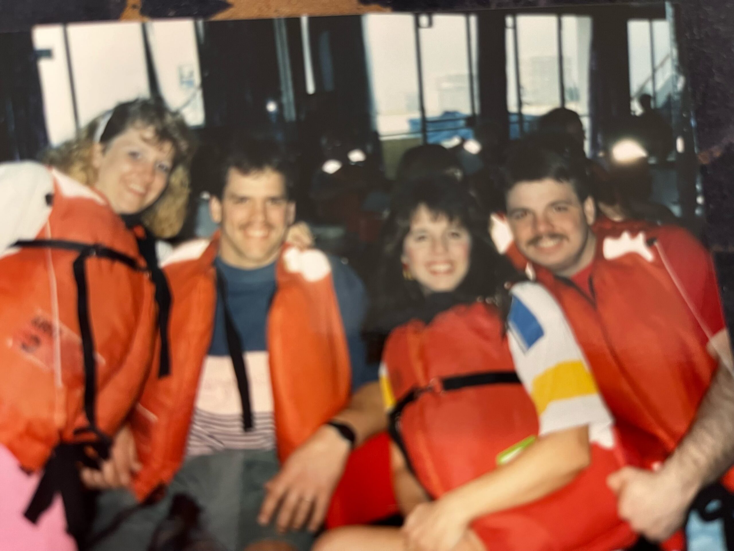 Group of people in orange life jackets posing for a photo indoors, likely on a boat or at a marine facility, smiling at the camera.