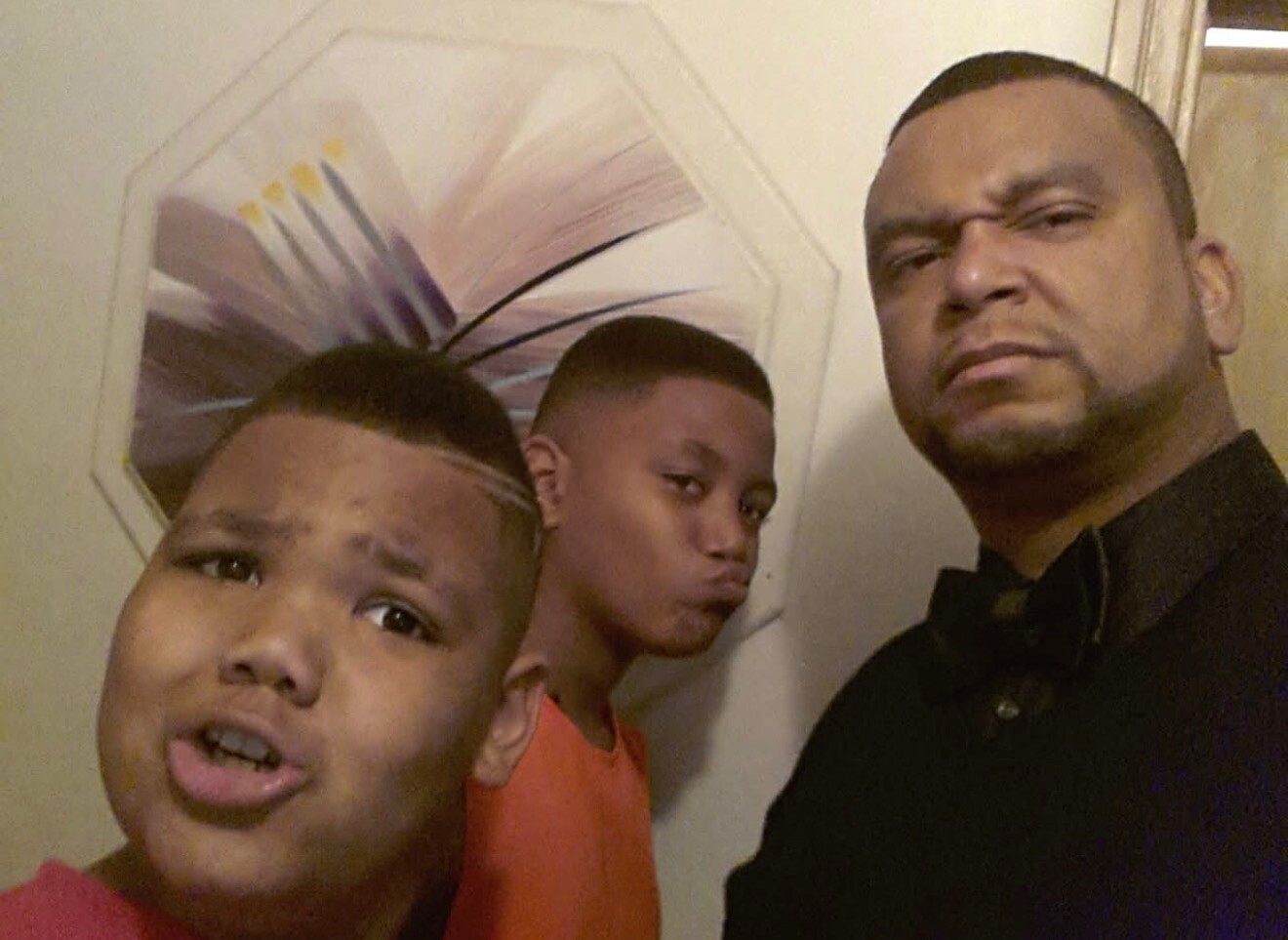 Three people posing for a selfie: a man in a dark shirt on the right and two boys in front left, one making a surprised face.