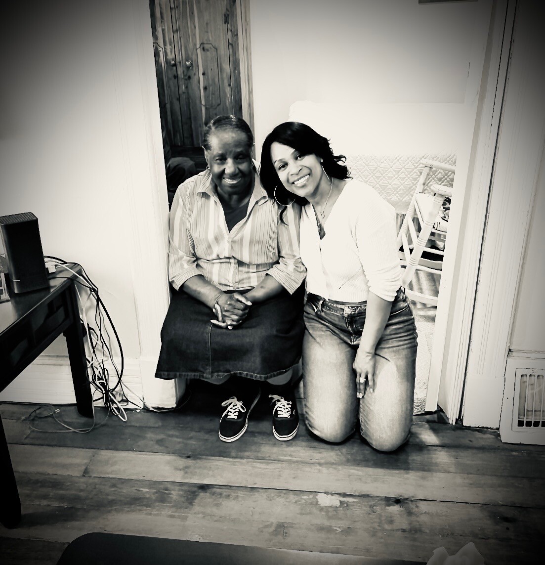 Two women kneeling in a doorway, smiling at the camera in a home interior; one wearing a striped shirt and denim skirt, the other in a white top and jeans.