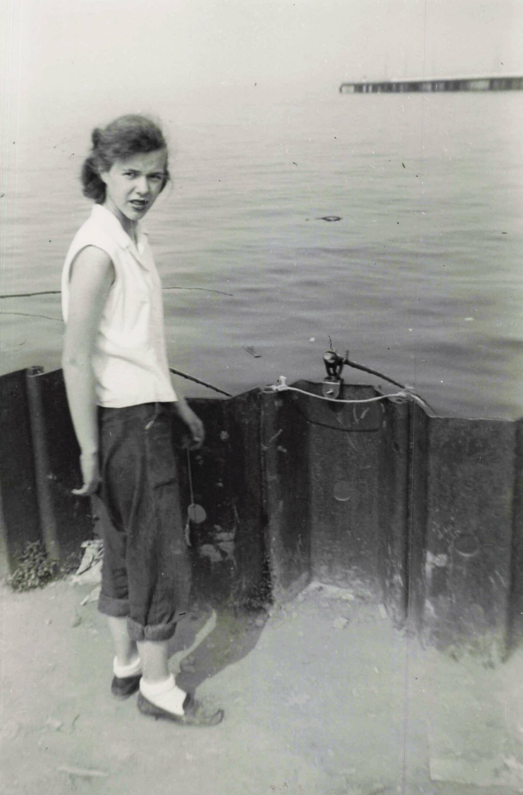 Girl standing on a dock near calm water, looking at the camera, wearing a sleeveless white top and rolled-up pants.