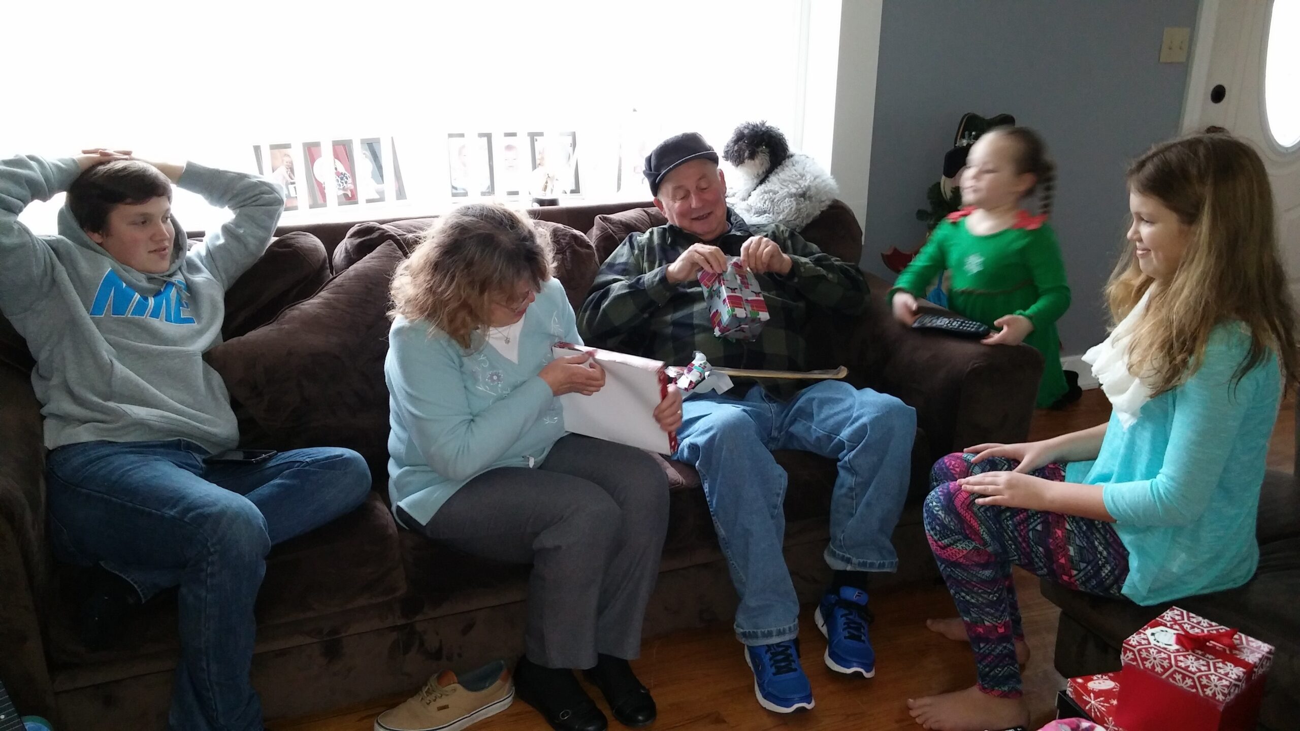 Family seated on a brown sofa opening Christmas gifts together indoors. Backlit window and festive wrapping visible.